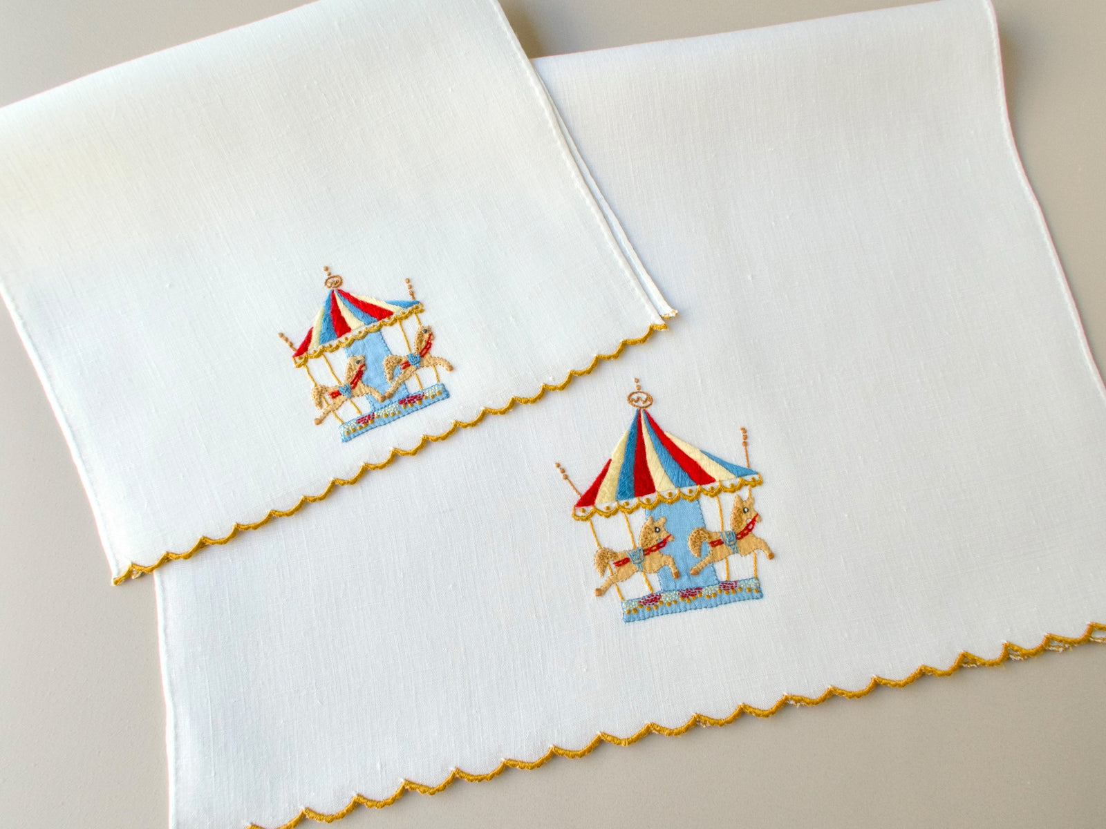 Vintage Madeira linen fingertip towel stacked on hand towel, both showing hand embroidered carousel design and scalloped gold hem, with the slight difference in linen shade visible between the two pieces​​​​​​​​​​​​​​​​