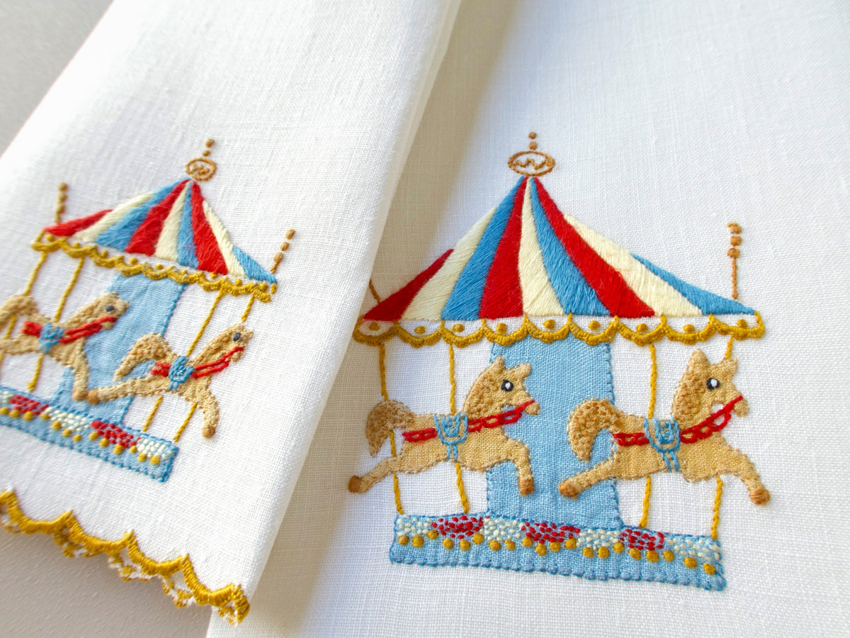 Close-up of vintage Madeira linen hand towel showing hand embroidered carousel with red, white and blue striped tent, golden horses and gilded poles on a light blue platform, with scalloped gold hem