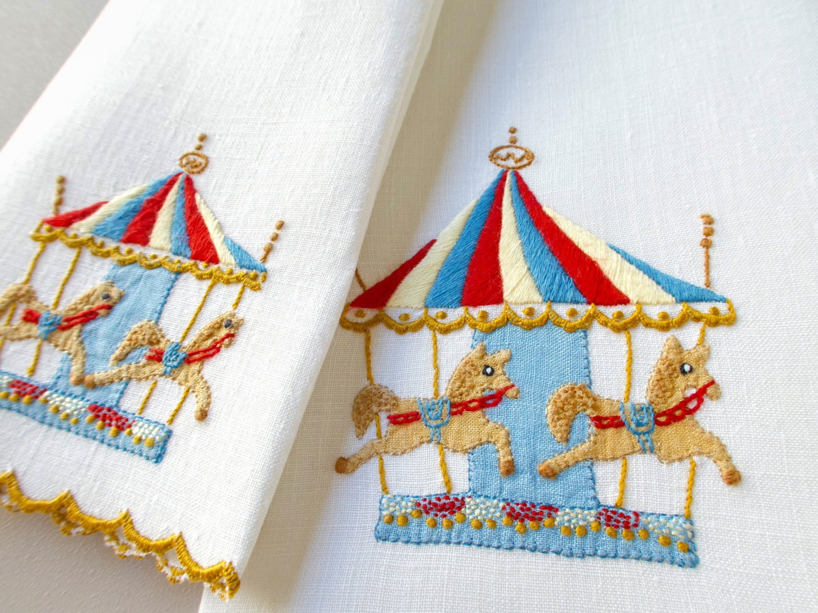 Close-up of vintage Madeira linen hand towel showing hand embroidered carousel with red, white and blue striped tent, golden horses and gilded poles on a light blue platform, with scalloped gold hem