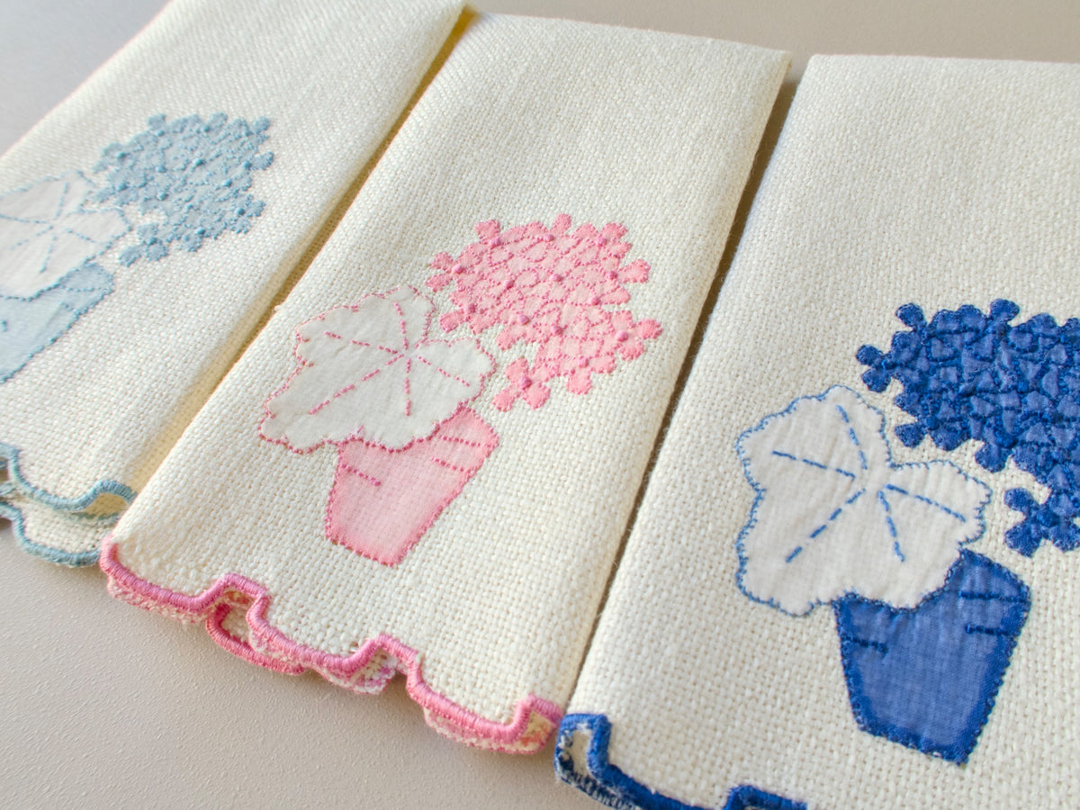 Three vintage Marghab marcasual linen fingertip towels fanned to show the “Geranium” appliqué design in powder blue, pink and cobalt, each with a matching stepped hem