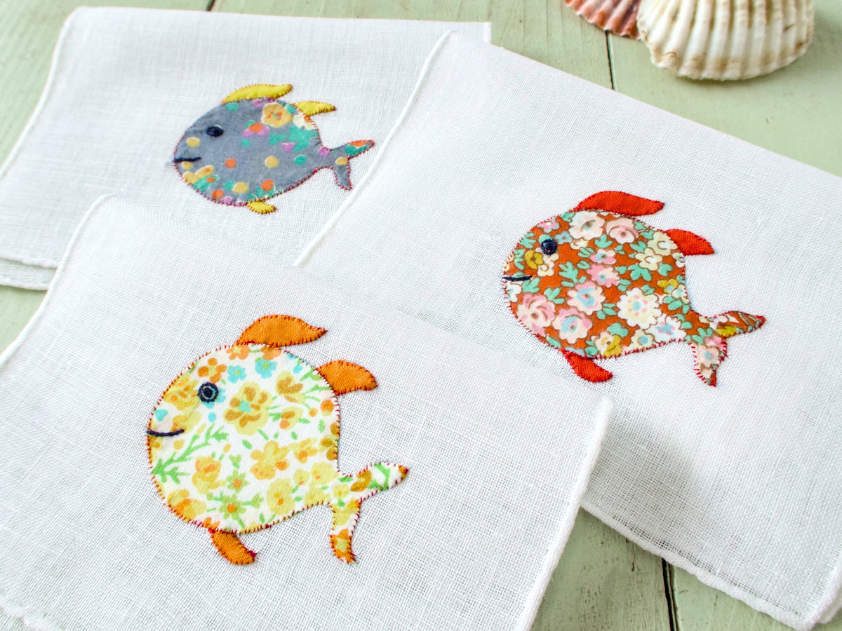 Three vintage Italian cocktail napkins fanned out, white semi-sheer linen, cheerful floral appliqué fish in different colorful prints