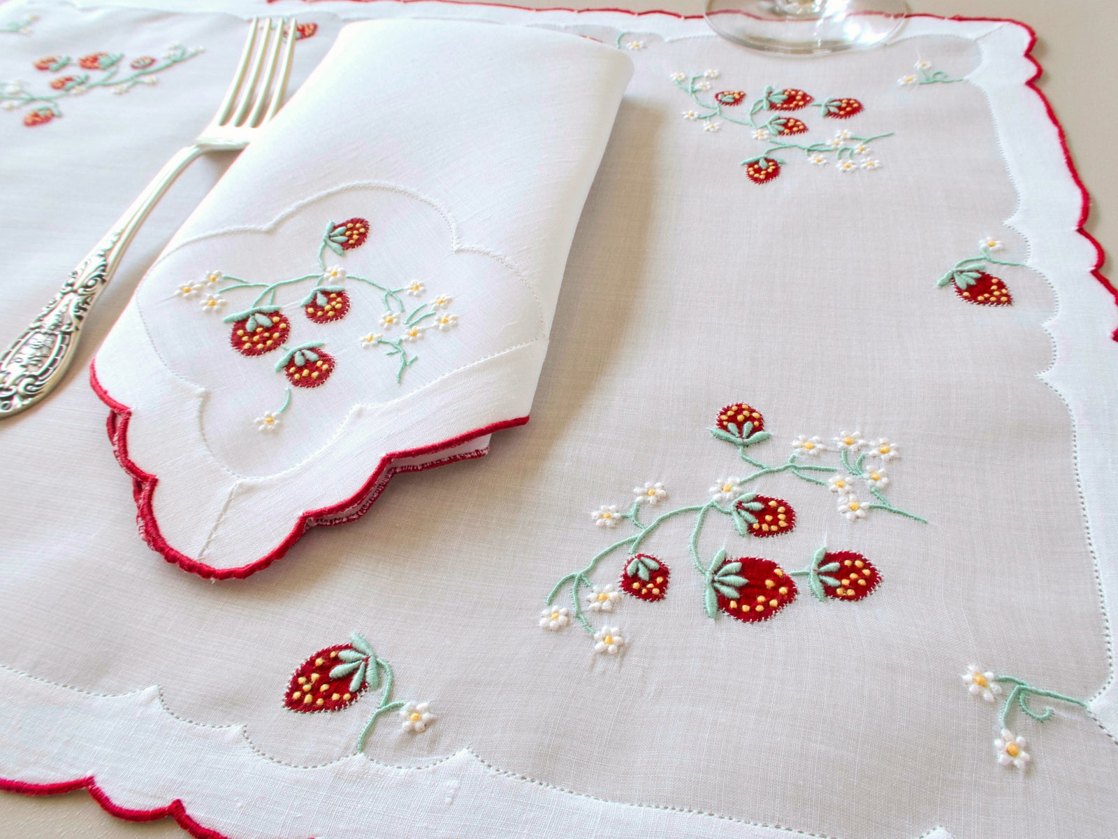 Vintage Madeira organdy placemat with folded linen napkin on top, red strawberry and white daisy embroidery with red scalloped border, fork and wine glass alongside