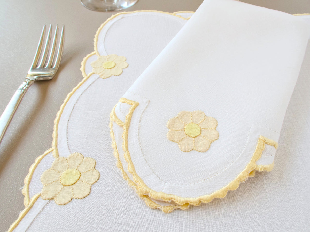 Vintage Madeira placemat at an angle with folded napkin on top, creamy Irish linen with daisy appliqué and yellow caseado bastido scalloped border, fork alongside