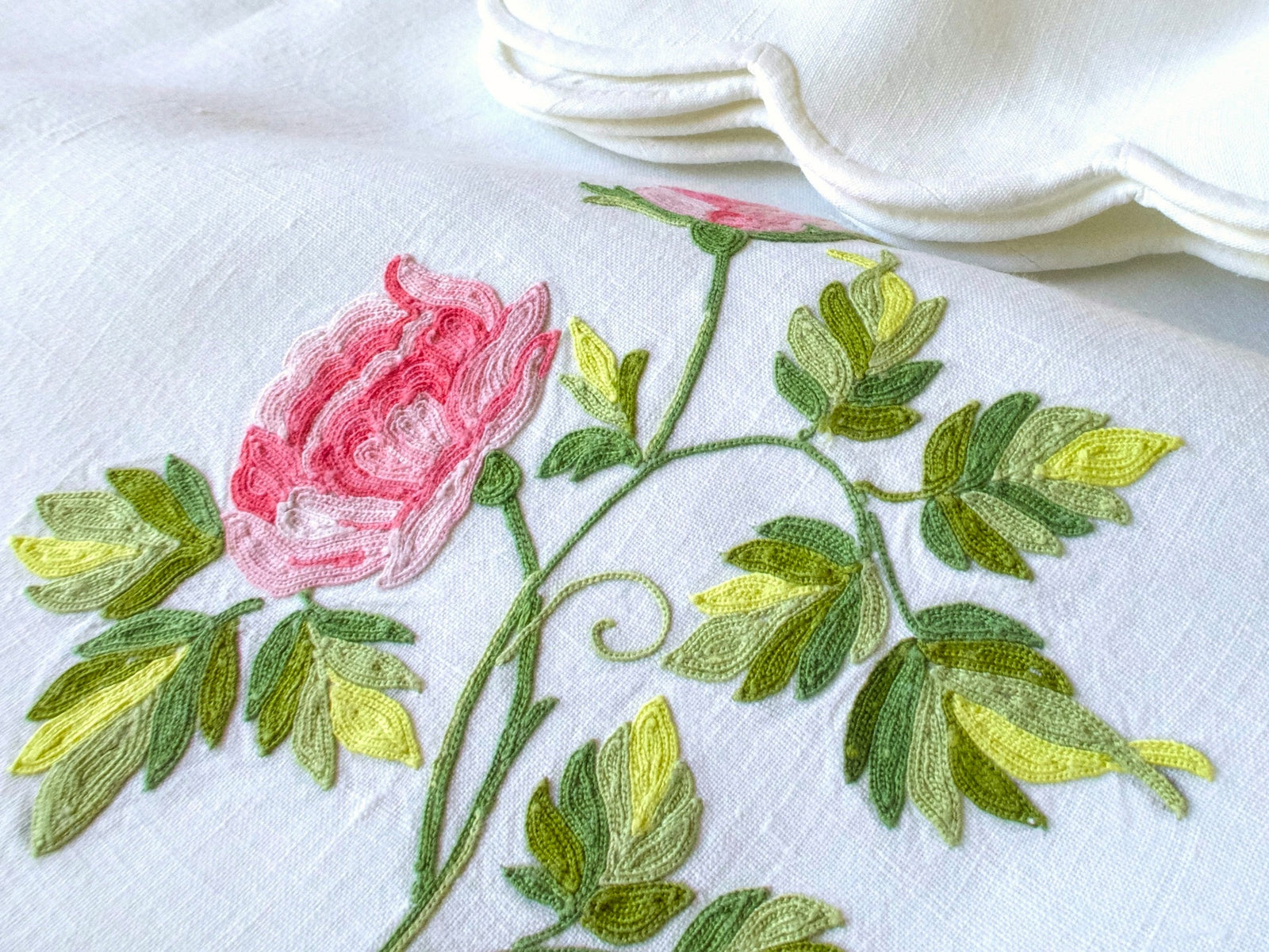 Close-up of Point de Beauvais embroidered pink rose in coral and blush threads with scrolling green stems and yellow-green leaves on white linen