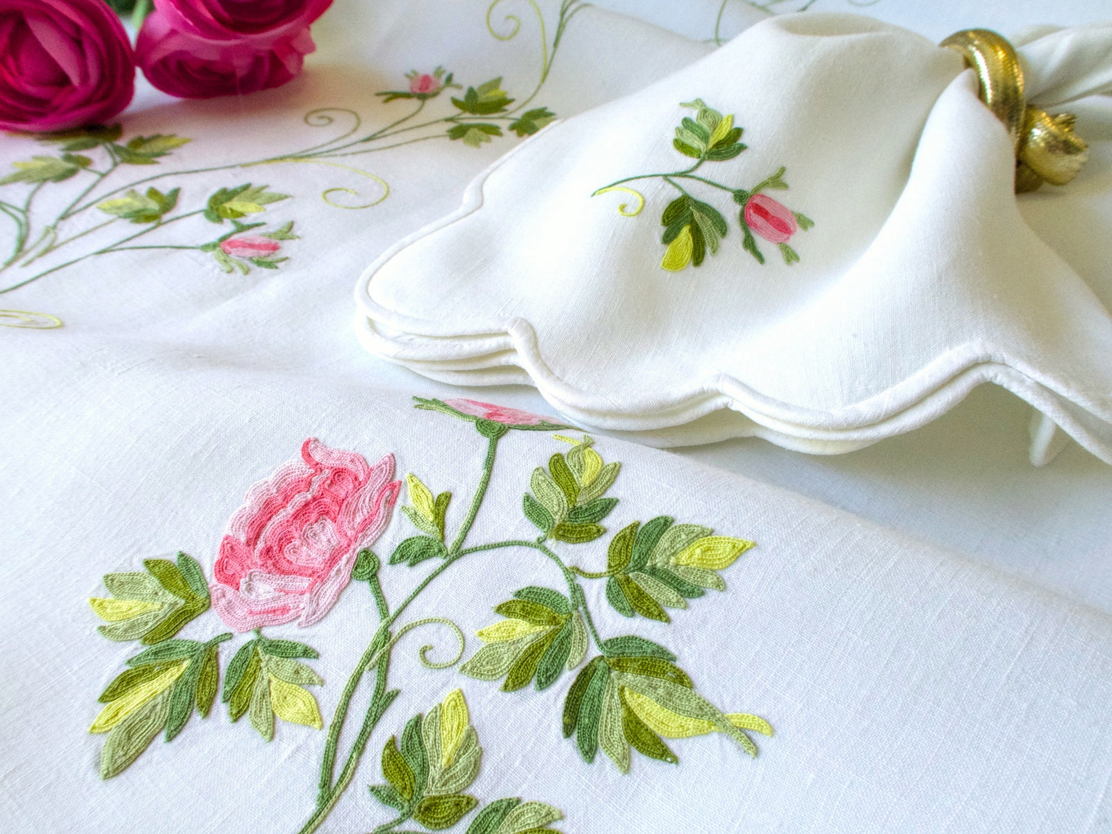 D. Porthault tablecloth with vining rose embroidery alongside scalloped napkins stacked with gold napkin ring, fresh pink roses in background