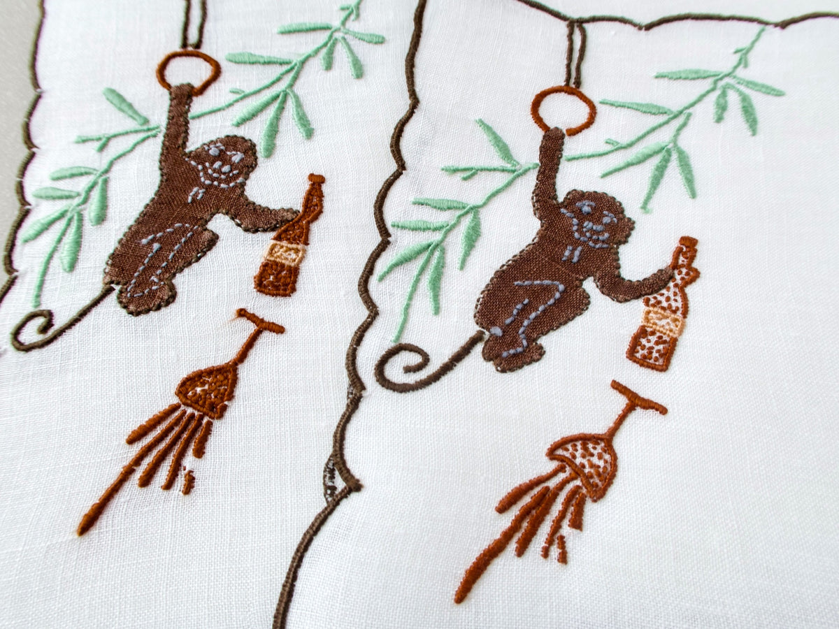 Two vintage Madeira cocktail napkins side by side, white linen with dark scalloped border, brown appliqué monkeys swinging from rings with champagne bottles and spilled glasses below
