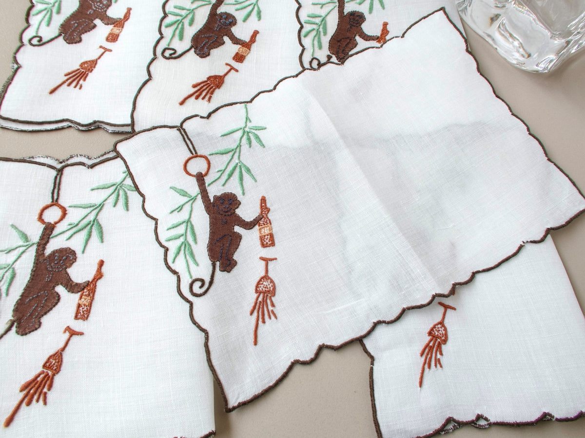 Single vintage Madeira cocktail napkin open and flat, brown appliqué monkey swinging from ring with bottle and spilled glass, green bamboo leaves above