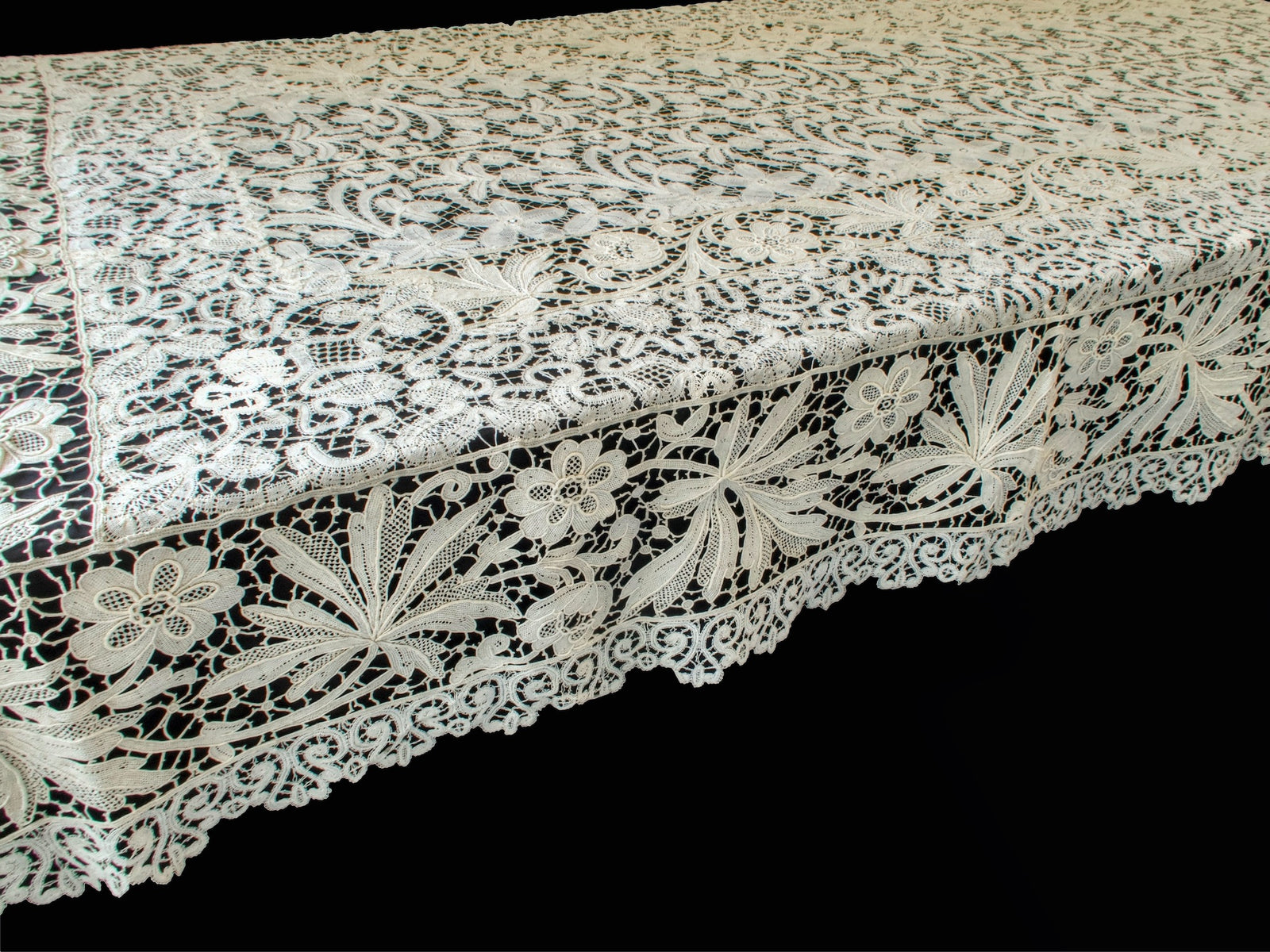 Close view of the deeply scalloped scrolling border with fan-shaped leaves and open flowers on an antique French lace tablecloth