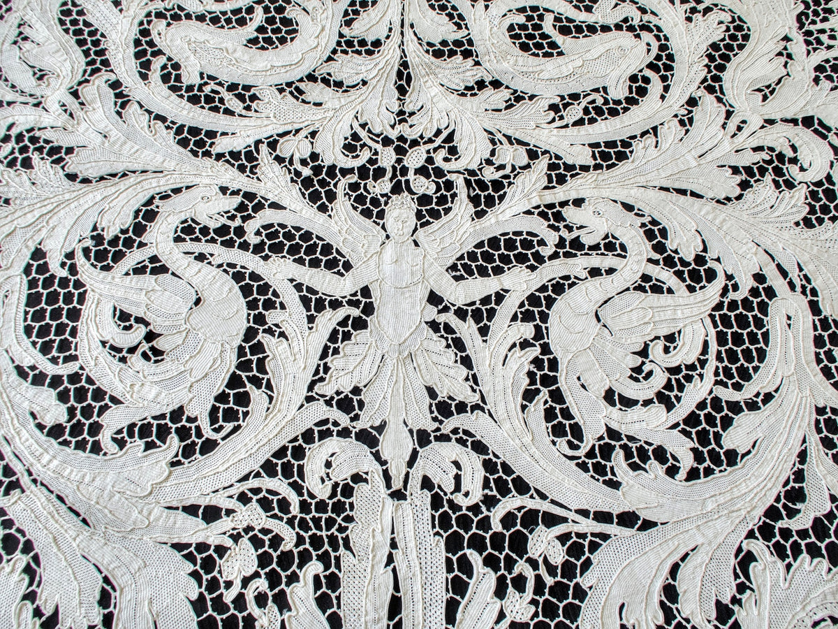 Winged figure flanked by dragonlike creatures and elaborate foliage in the lace.