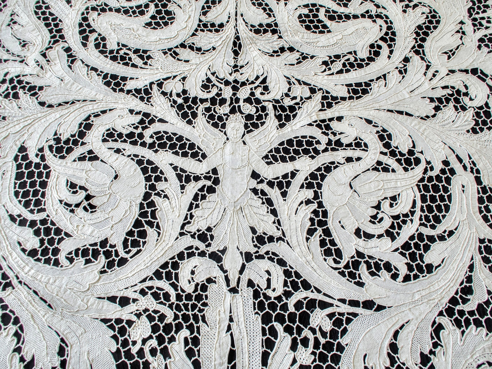 Winged figure flanked by dragonlike creatures and elaborate foliage in the lace.