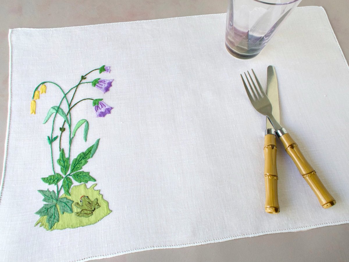 Vintage Italian linen placemat showing hand embroidered frog among flowers and leaves, with fork, knife and cup for scale