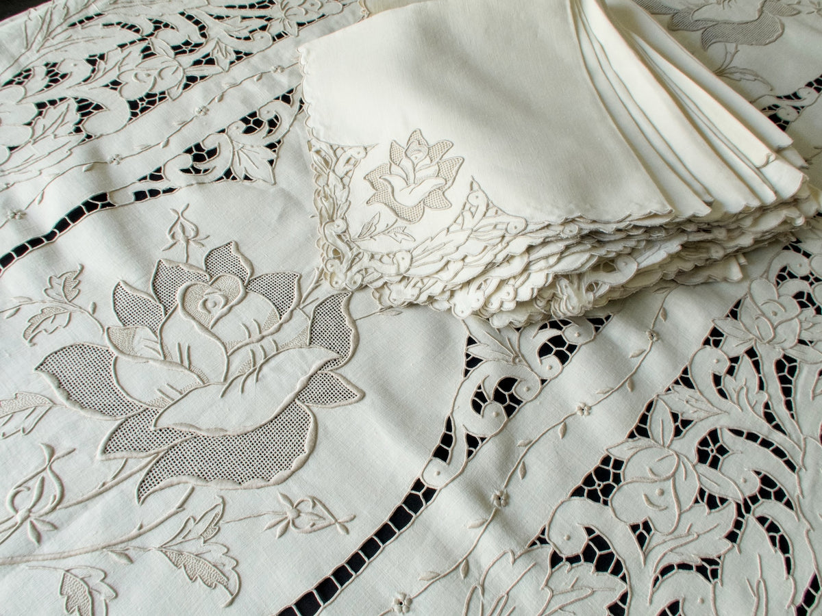 Stack of matching napkins and tablecloth corner showing sculptural rose embroidery on linen.