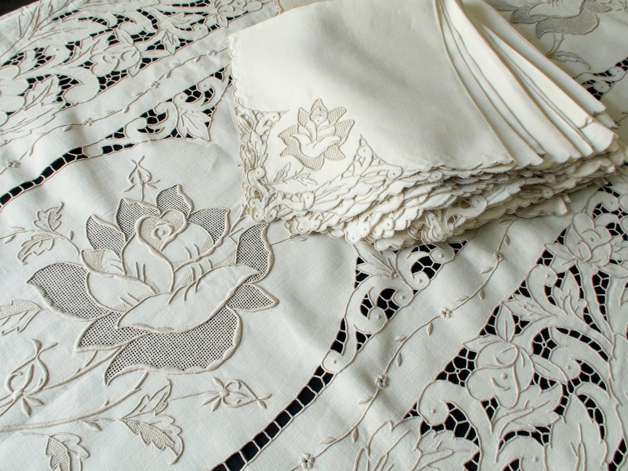 Stack of matching napkins and tablecloth corner showing sculptural rose embroidery on linen.