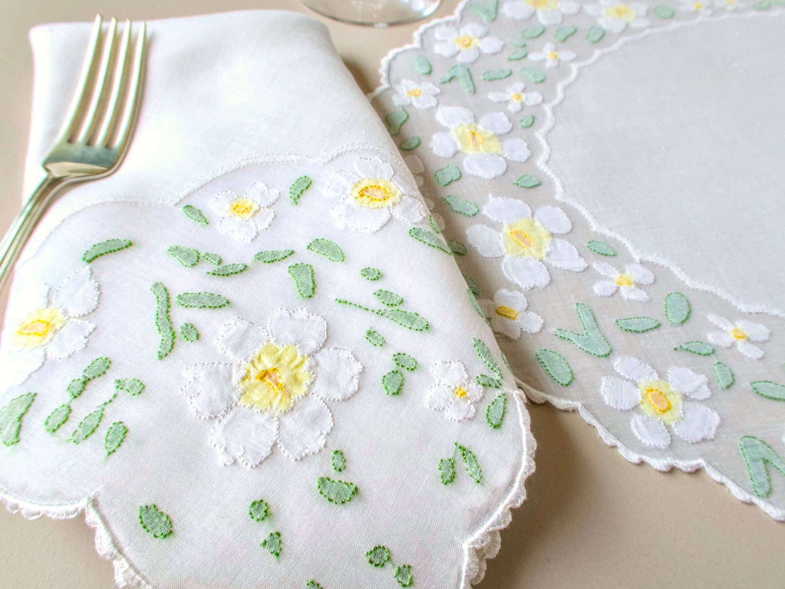 Folded daisy-appliquéd napkin and oval Madeira organdy placemat showing scalloped edge and white daisy detail on warm beige background