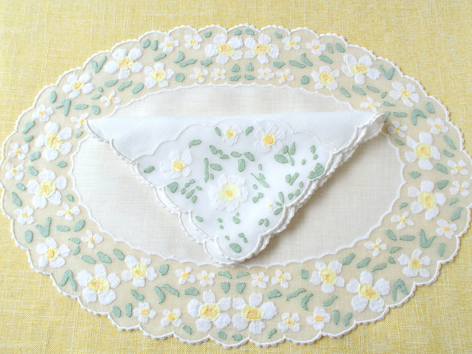 Overhead view of oval vintage Madeira organdy placemat and matching napkin with white daisy appliqué and scalloped border on yellow linen tablecloth