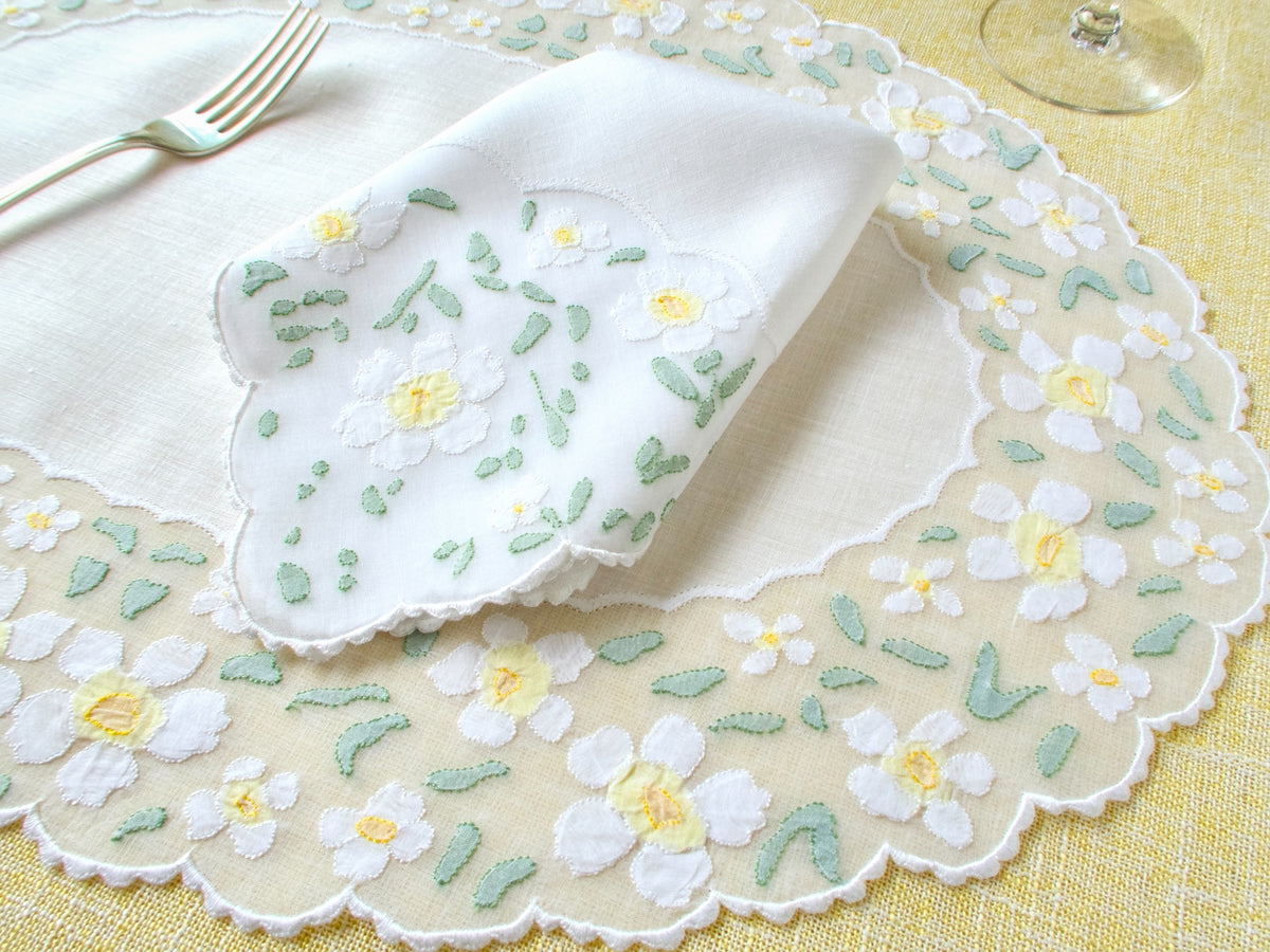 Oval vintage Madeira organdy placemat with white appliquéd daisies, yellow centers and sage green leaves, styled with folded napkin and silver fork on yellow linen