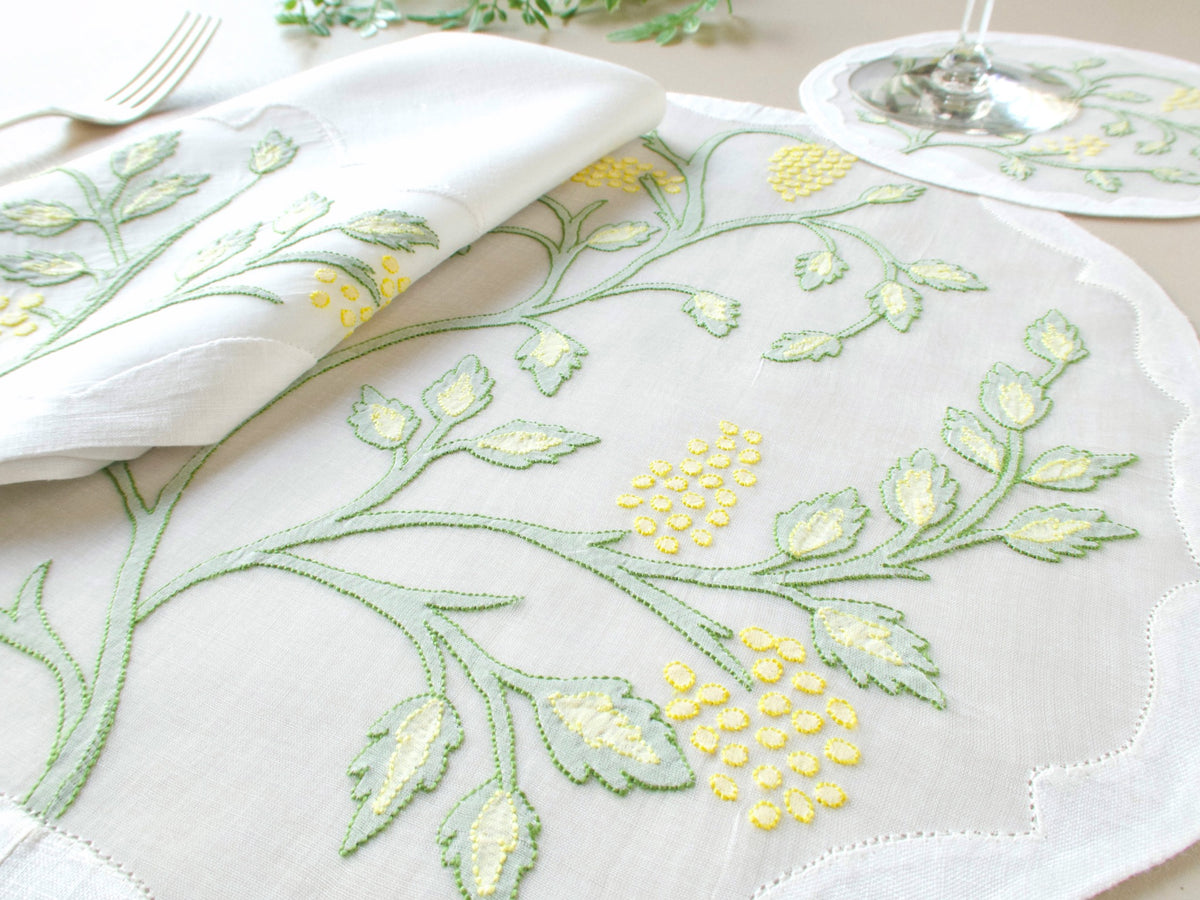 Angled close-up of vintage Madeira organdy placemat with Jacobean botanical shadow work embroidery in sage green and yellow, napkin and coaster visible in background