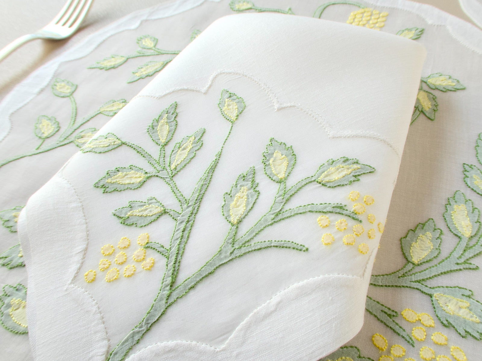 Close-up of linen napkin showing Jacobean botanical shadow work embroidery in sage green and yellow on organdy placemat