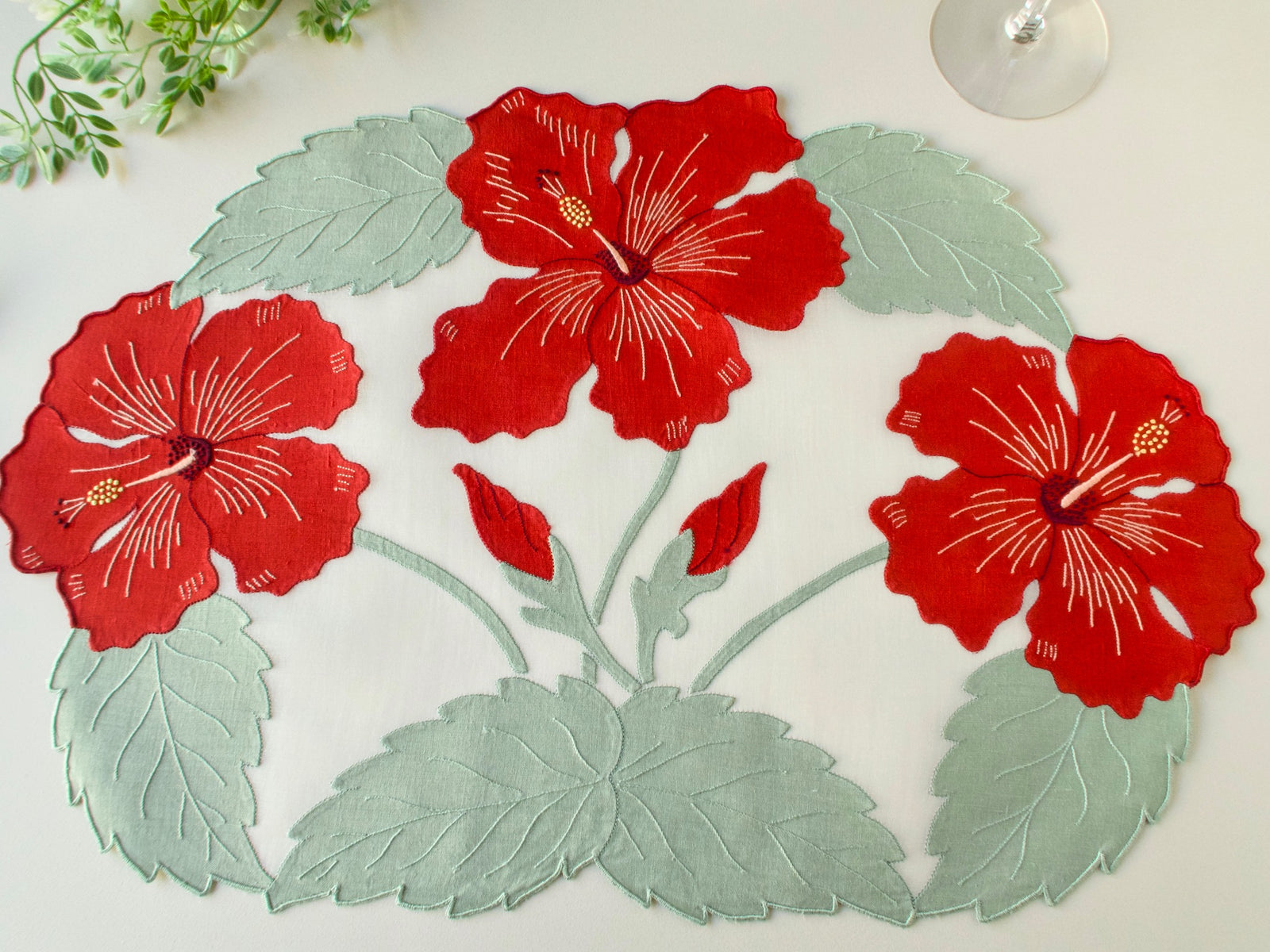 Single vintage Madeira organdy placemat showing full linen appliqué design of three red hibiscus blooms, buds and sage green leaves