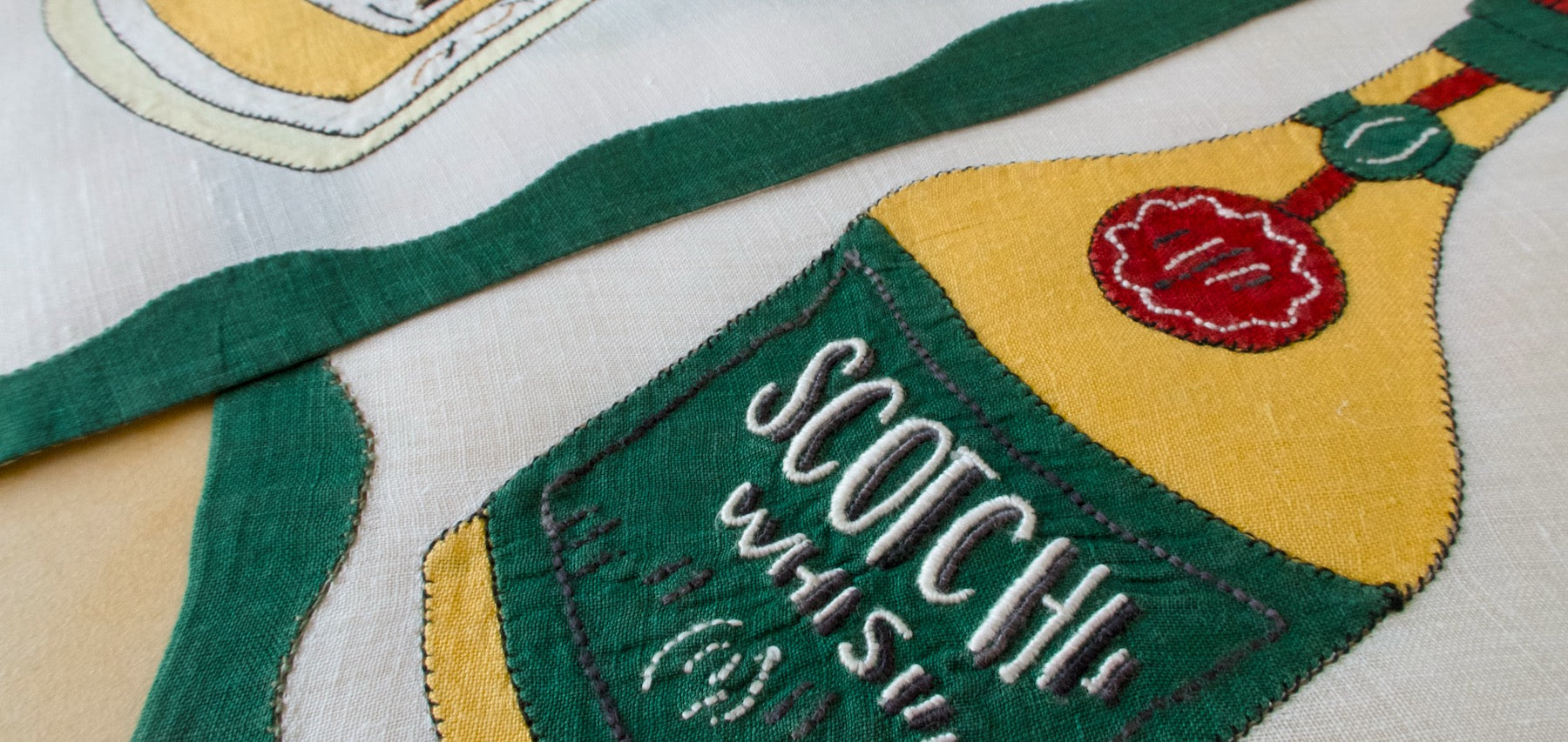 Embroidered design of a bottle labeled 'Gin' and another labeled 'Scotch Whisky' on fabric, vintage cocktail napkins from Madeira