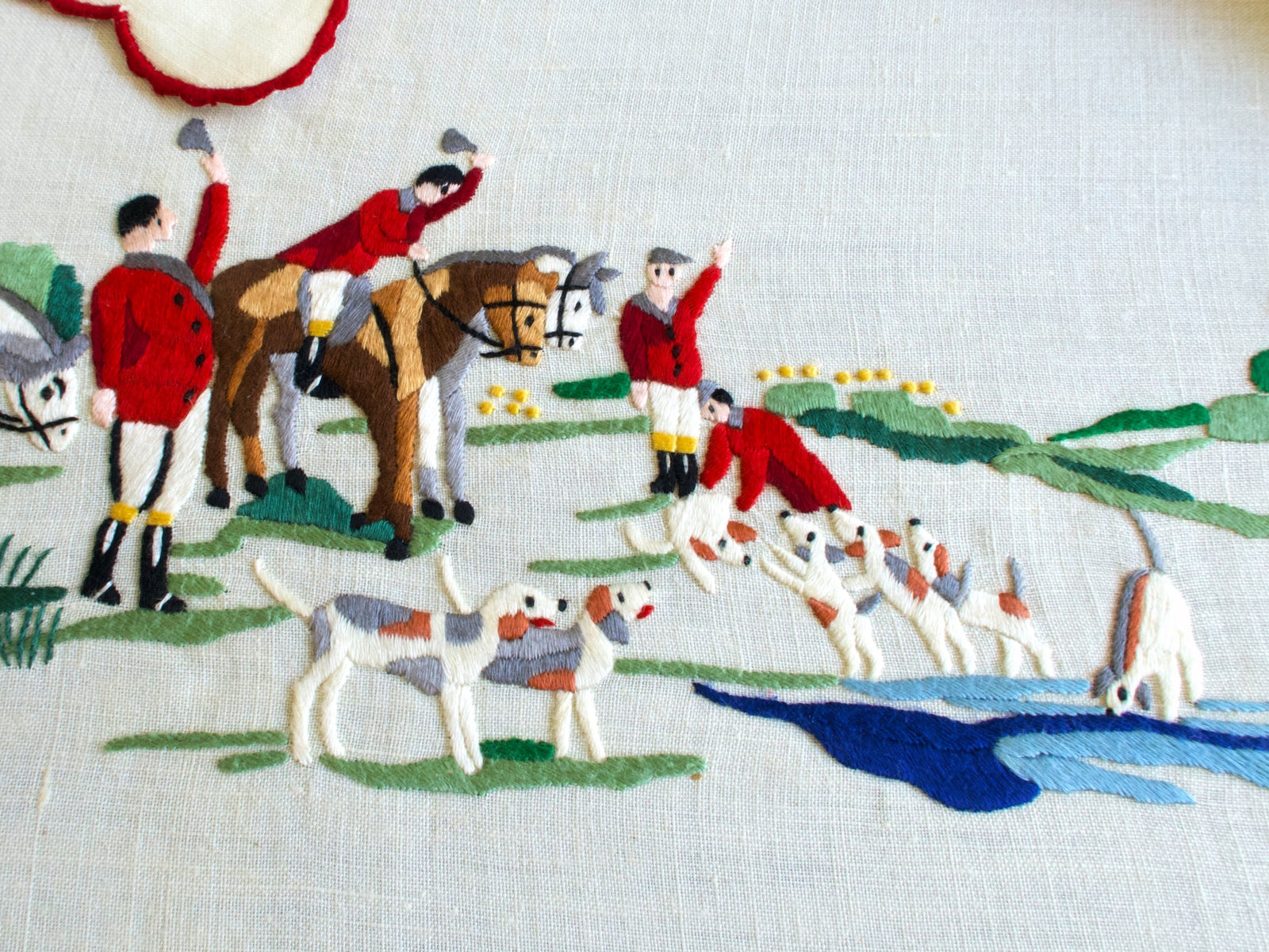 Hand embroidered huntsmen waving caps beside a blue stream, with hounds gathered at the water
