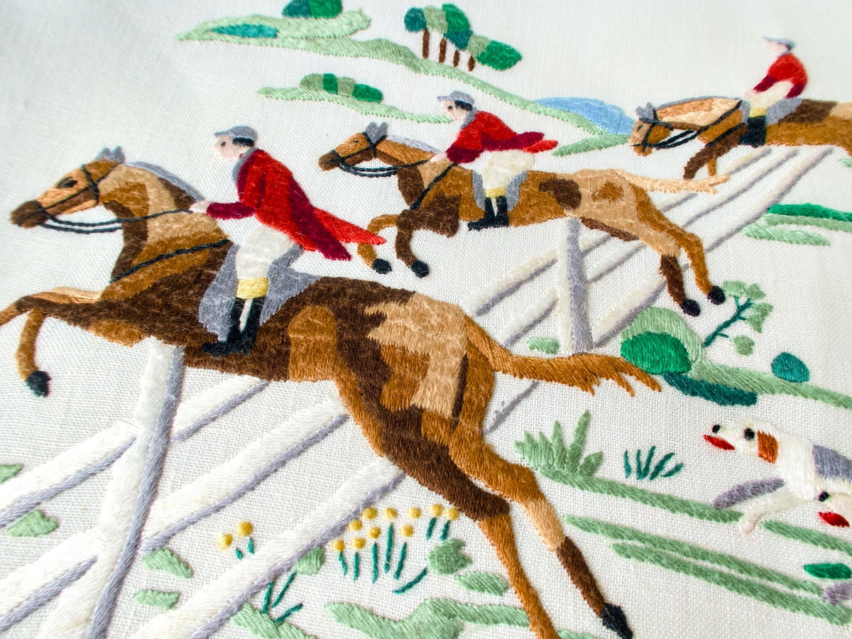 Vintage Madeira tablecloth with hand embroidered red-coated riders on galloping horses clearing a white fence, hounds racing below