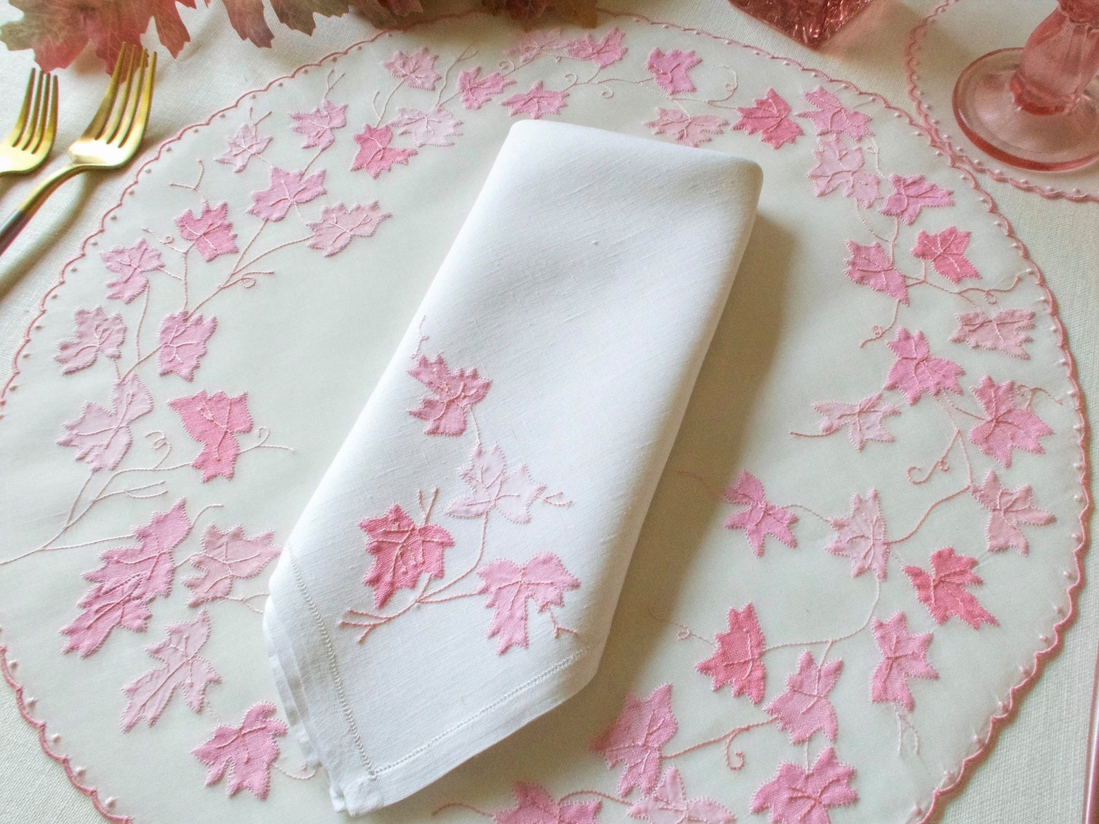 Overhead view of vintage hand embroidered organdy placemat with pink appliqué maple leaf design and matching linen napkin, pink glass on matching coaster in background