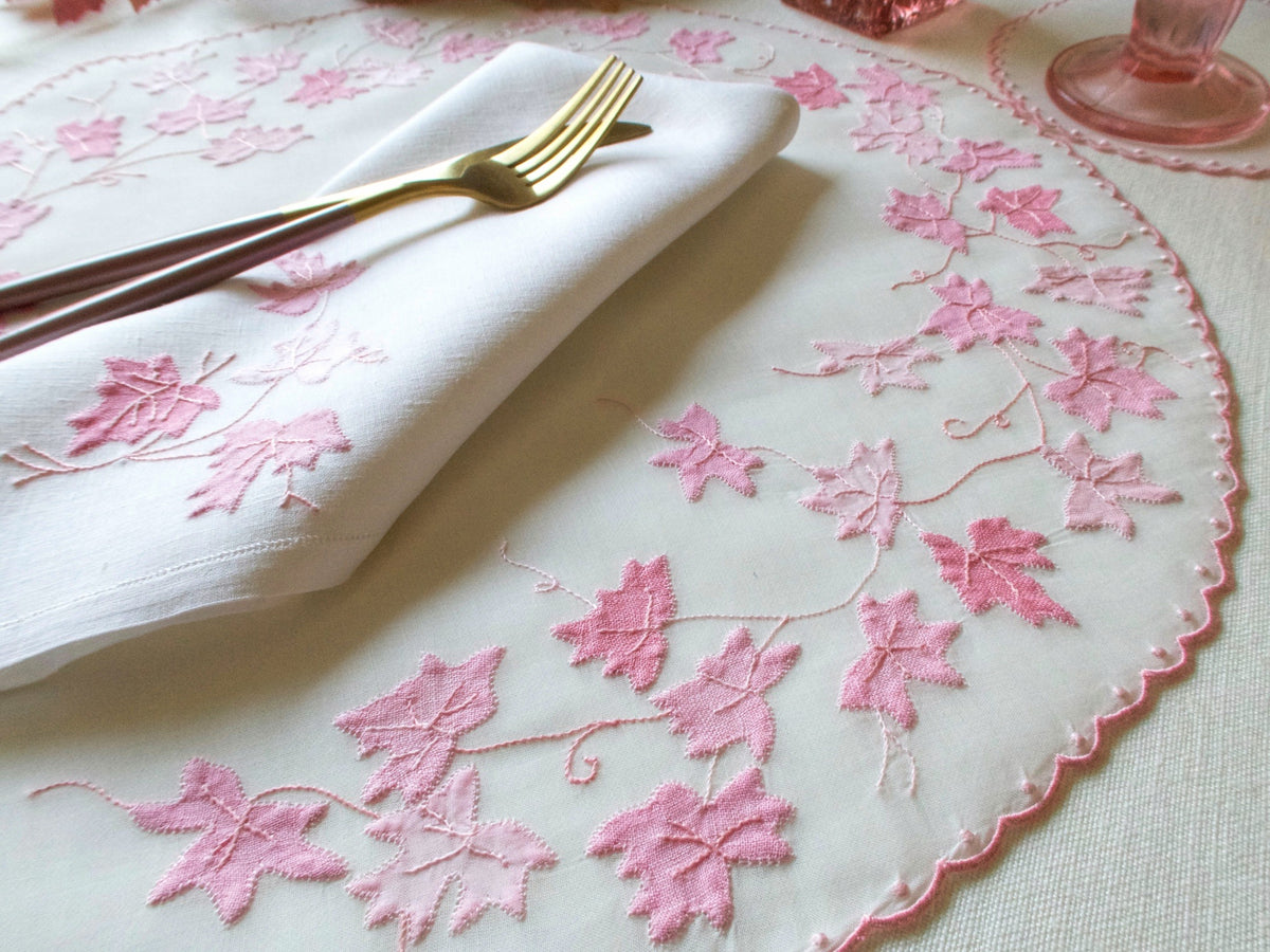 Angled close-up of vintage hand embroidered organdy placemat with pink appliqué leaf vines, linen napkin and gold cutlery on white tablecloth