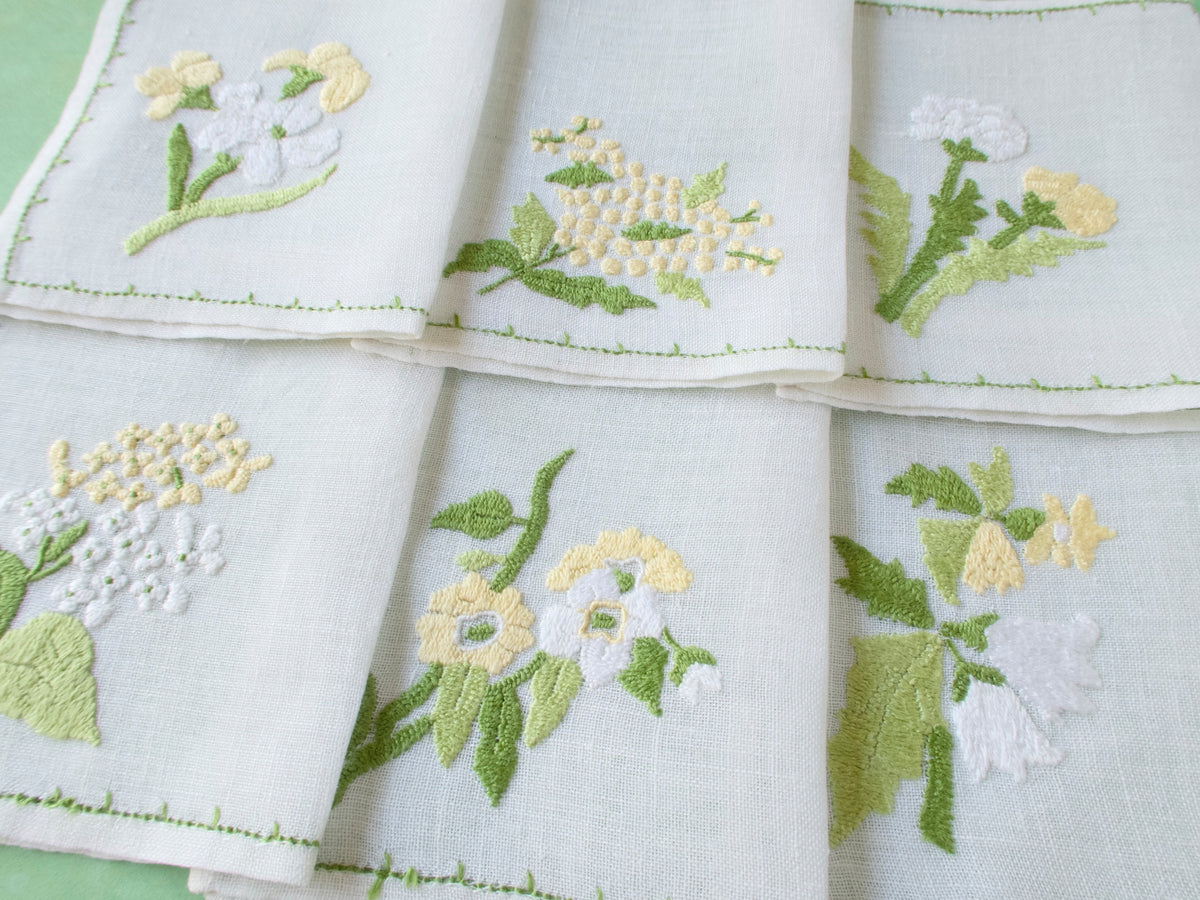 Close view of Rapisardi floral cocktail napkins showing dimensional hand embroidery and green stitched border on semi-sheer linen.