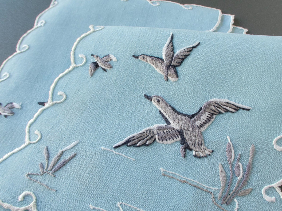 Pale blue Madeira cocktail napkins embroidered with ducks in flight above reeds, finished with white scrolling scalloped edges.