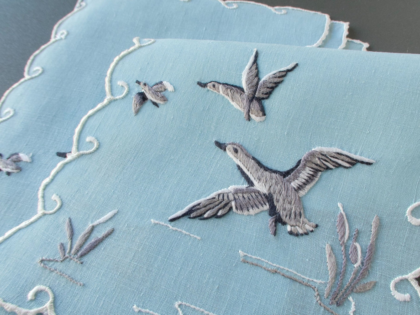 Pale blue Madeira cocktail napkins embroidered with ducks in flight above reeds, finished with white scrolling scalloped edges.