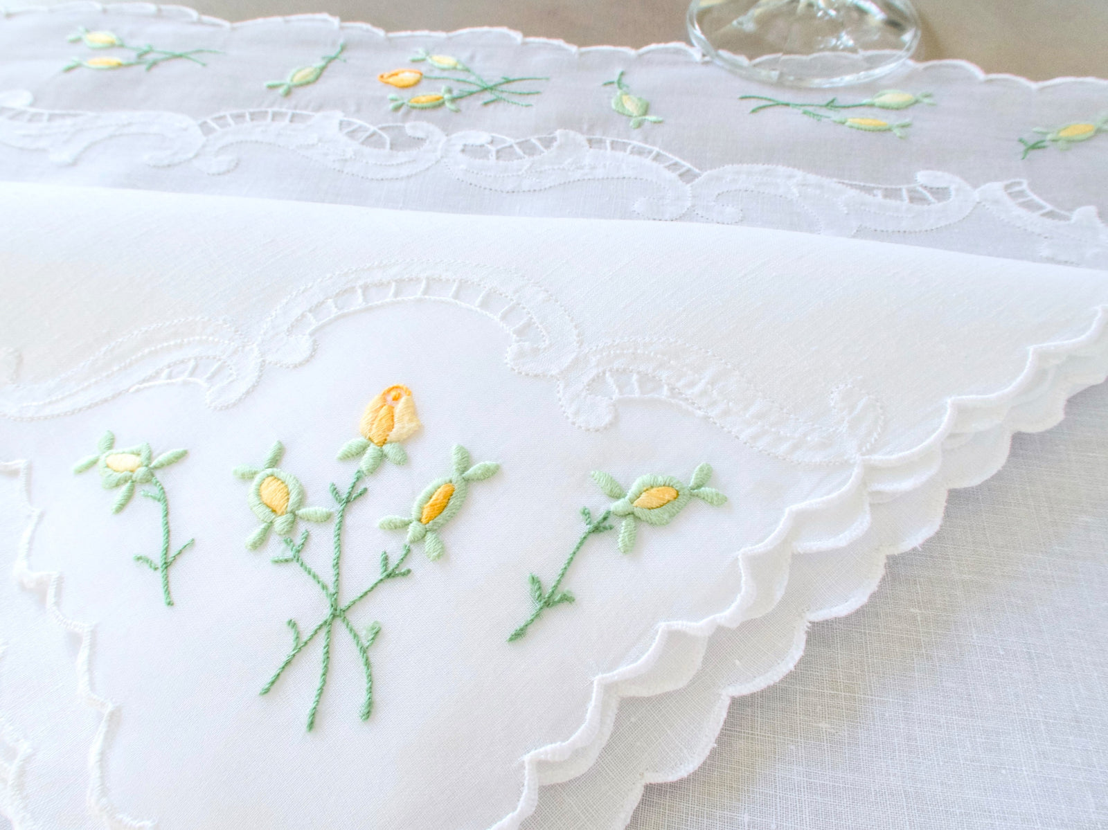 Vintage Madeira linen and organdy placemat with yellow rosebud embroidery on green stems and delicate scalloped edge, with wine glass in background