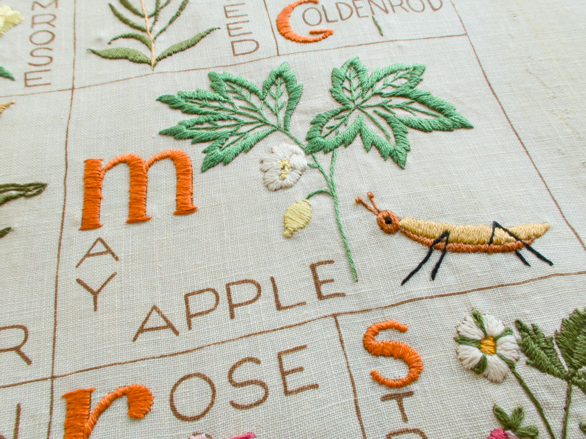 Vintage botanical alphabet sampler with May Apple plant and embroidered grasshopper detail on hand-embroidered linen.