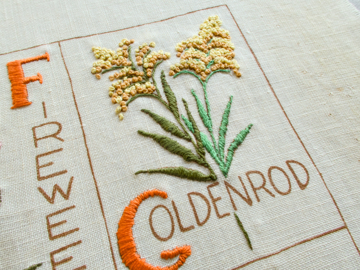 Detail of vintage botanical alphabet sampler showing embroidered goldenrod flower and lettering.