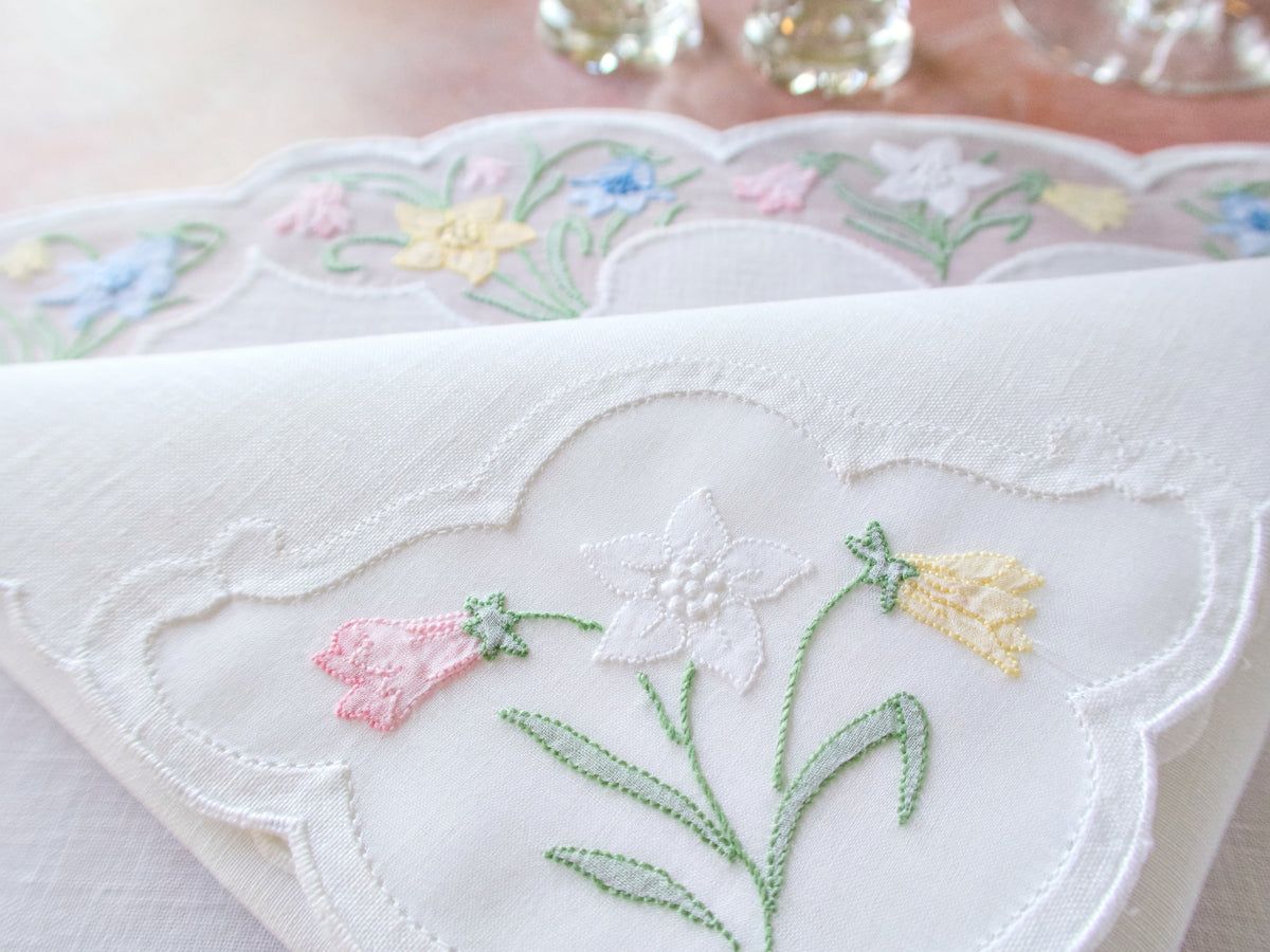 Close-up of vintage Madeira oval placemat showing daffodil and bluebell embroidery in organdy windows with scalloped edges on white linen.