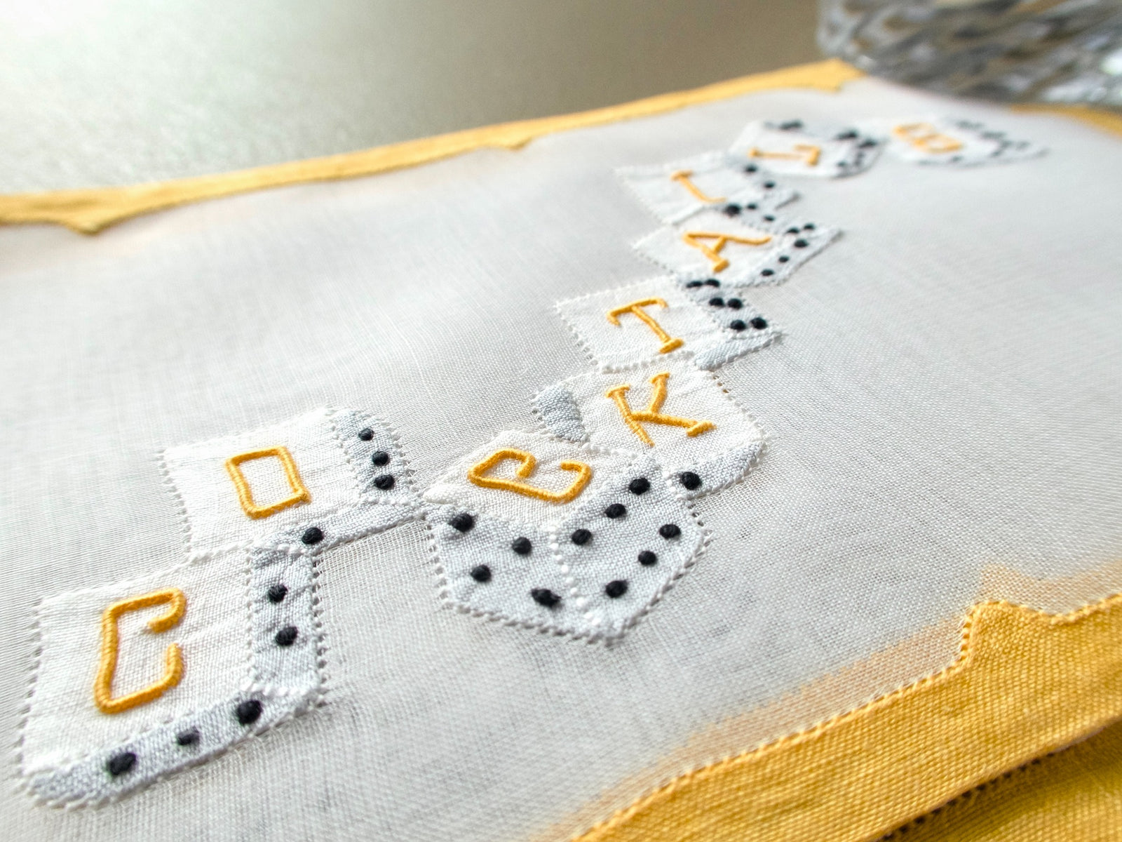 Close-up of white appliqué dice spelling “COCKTAILS” on sheer organdy Madeira cocktail napkins, with black dots, golden letters and yellow edging.
