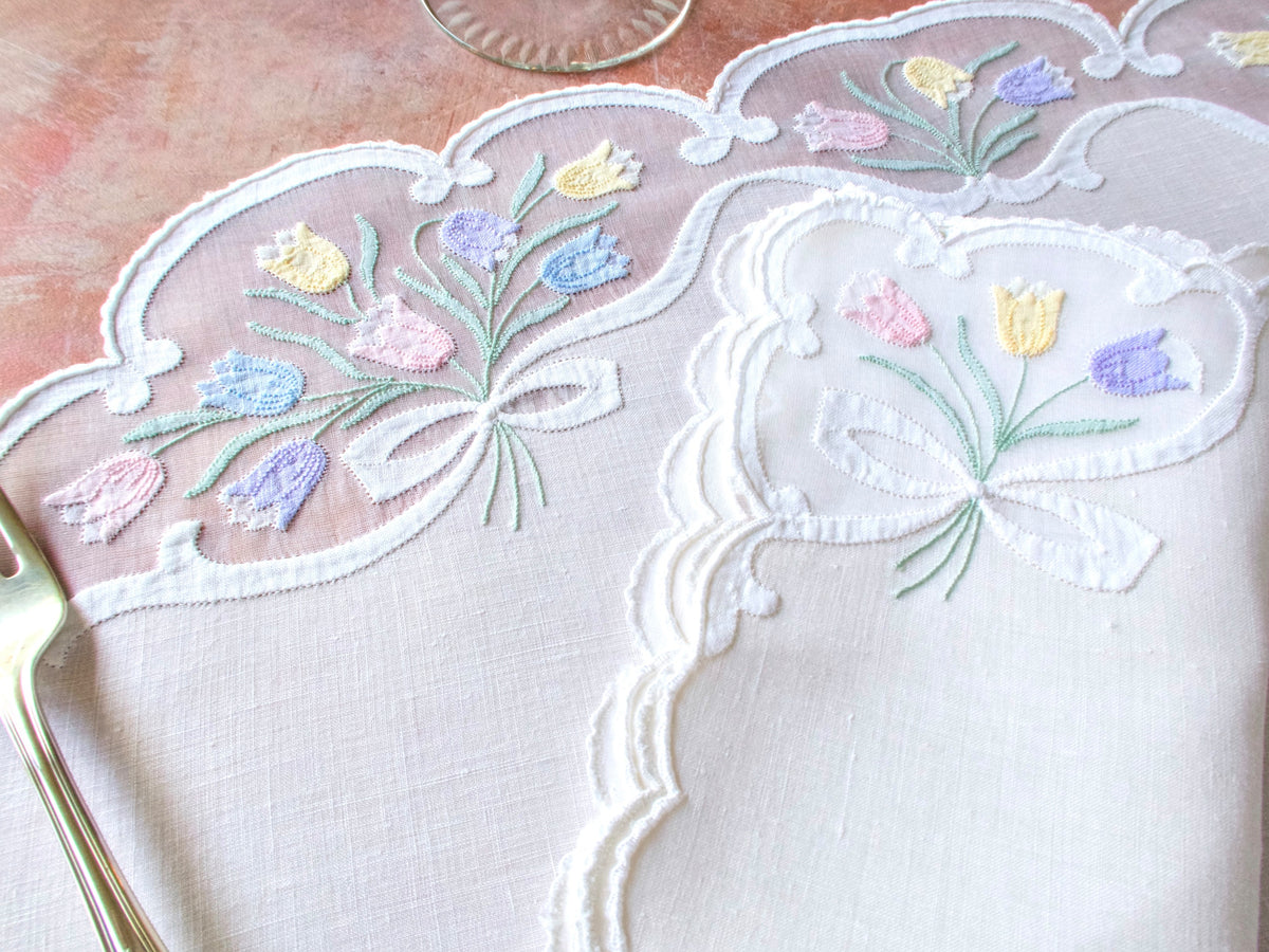 Close-up of vintage Madeira oval placemat showing tulip and bow embroidery in organdy windows with caseado bastido scalloped border on white linen.