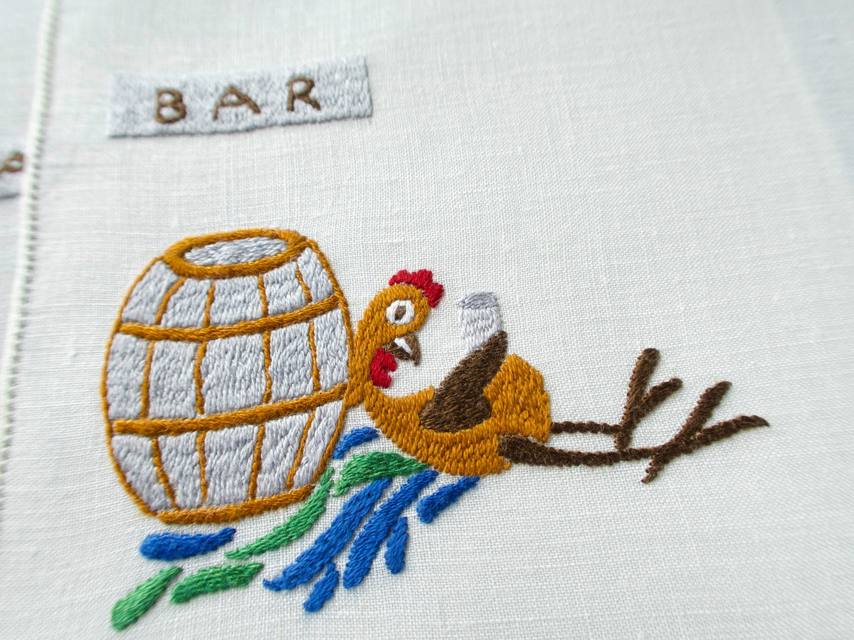 Close view of hand embroidered rooster beside a barrel on a vintage Rapisardi linen cocktail napkin
