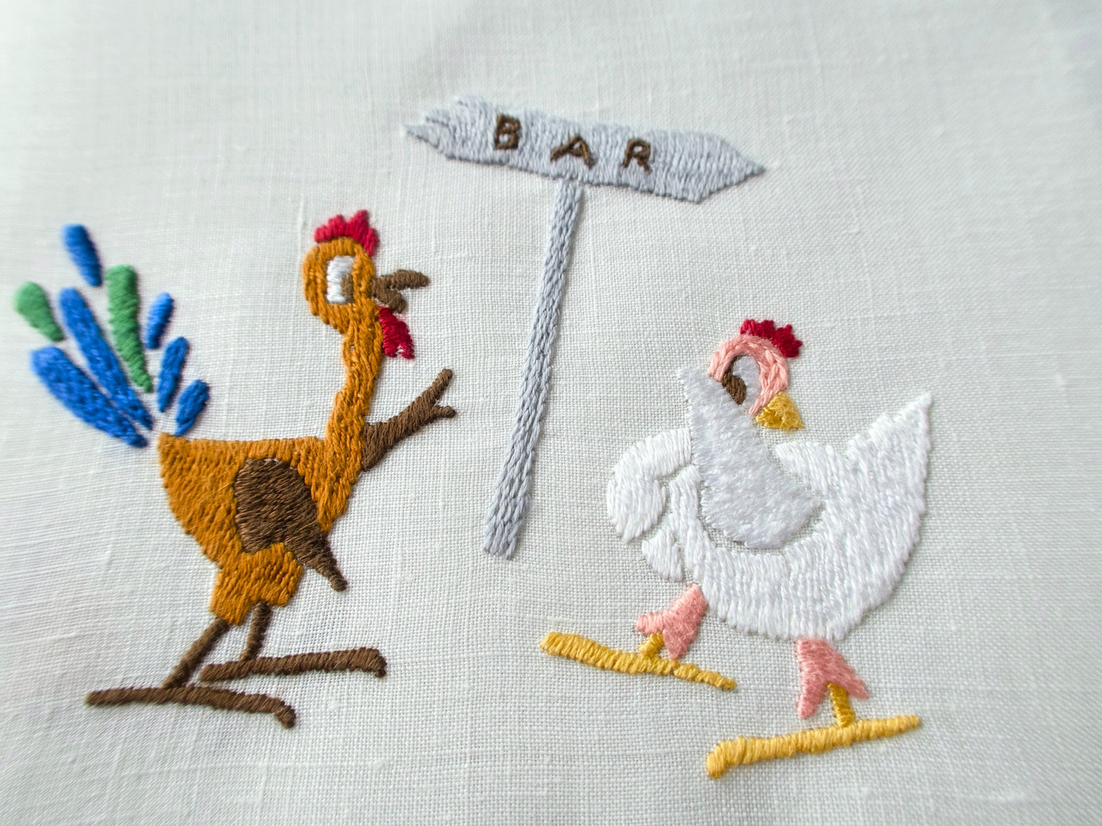 Close view of hand embroidered rooster and white hen flirting at a bar signpost on a vintage Rapisardi linen cocktail napkin