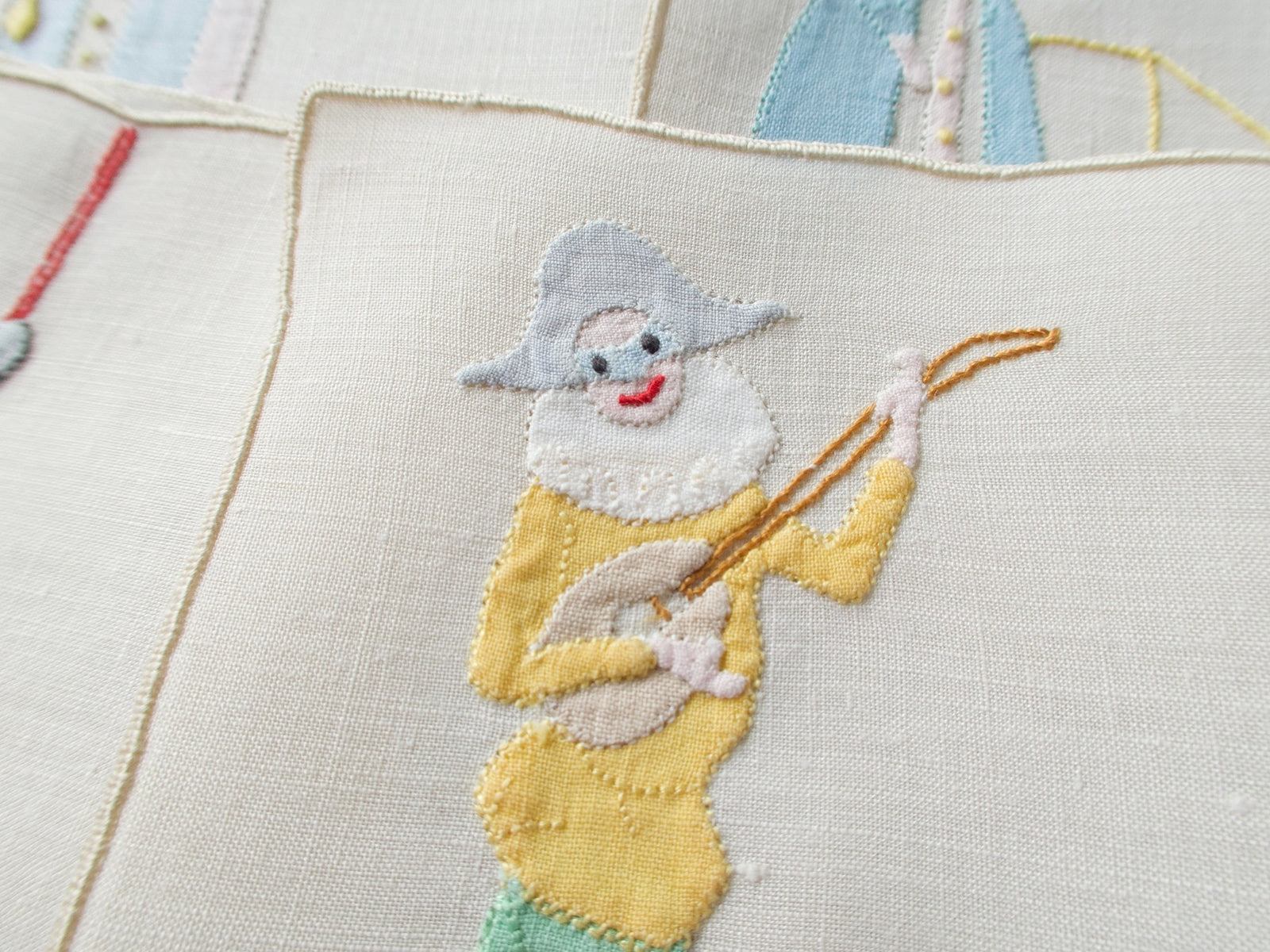 Close view of an appliqué joker in a gray hat and white ruff playing a lute on a vintage Madeira cocktail napkin