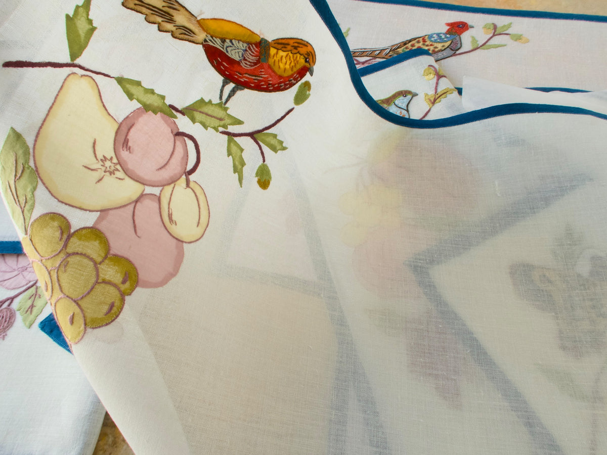 Semi-sheer off-white linen placemat held up to show the light passing through the fabric and the bird and fruit embroidery.