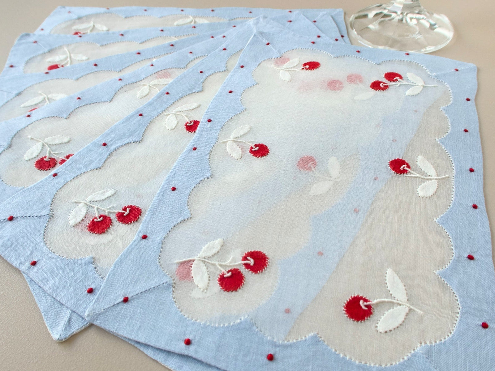 Six vintage Madeira cocktail napkins fanned out beside a wine glass, each with cherry appliqué on an organdy center inside a blue linen border
