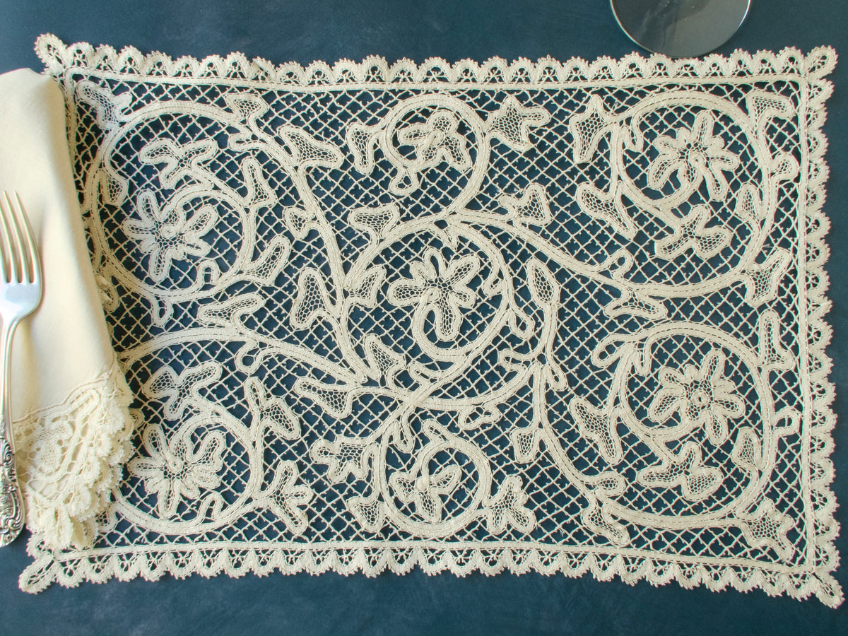 Vintage bobbin lace placemat with intricate scrolling floral motifs, styled with linen napkin and silver fork on dark background.