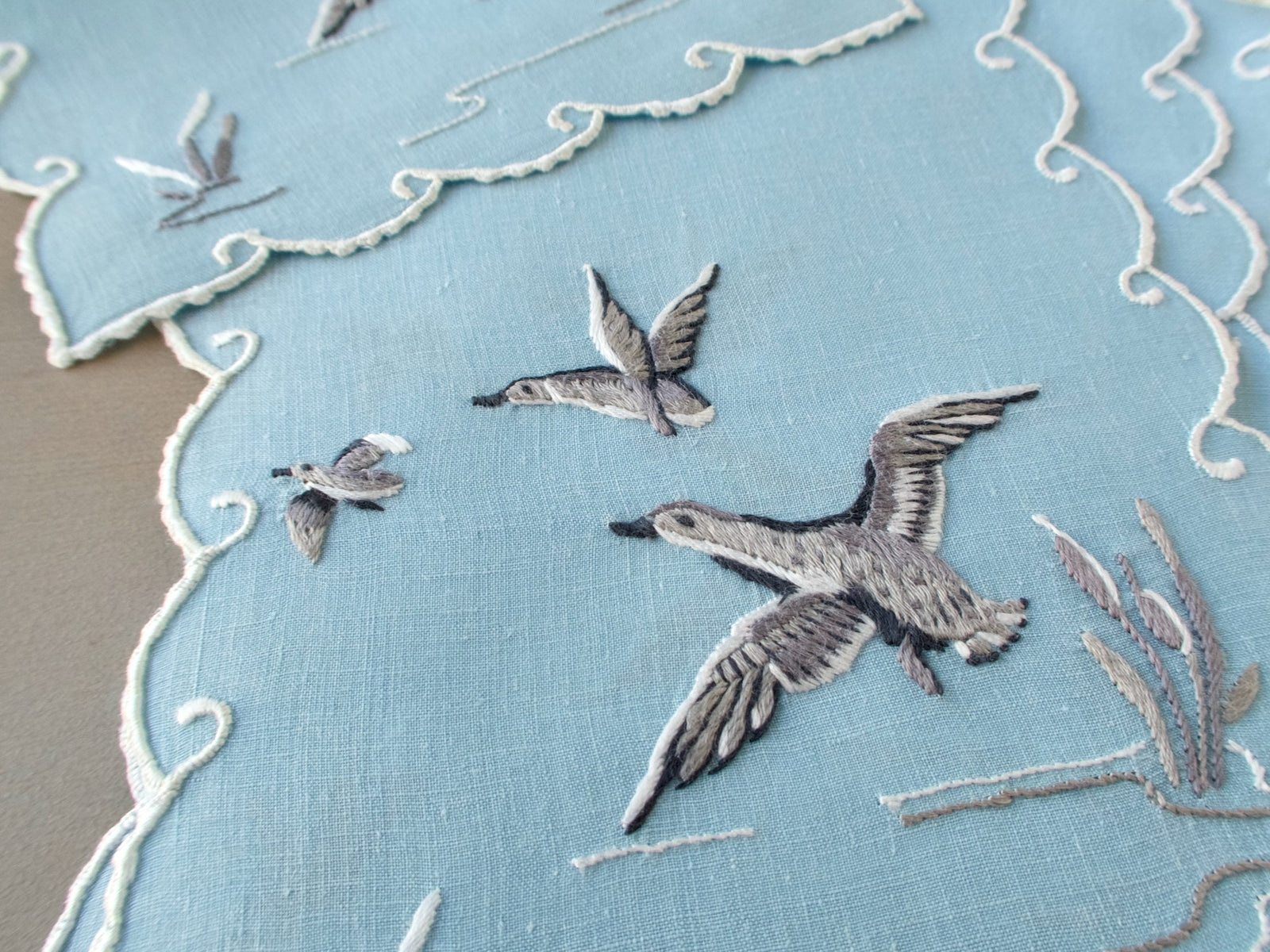 Close view of ducks in flight embroidered on pale blue linen, with white scrolling scalloped edges.