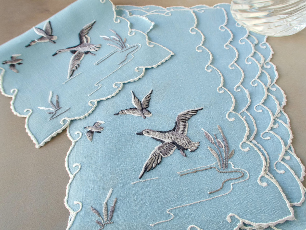 Six pale blue duck cocktail napkins arranged in a fan to show their embroidered birds and scalloped borders.