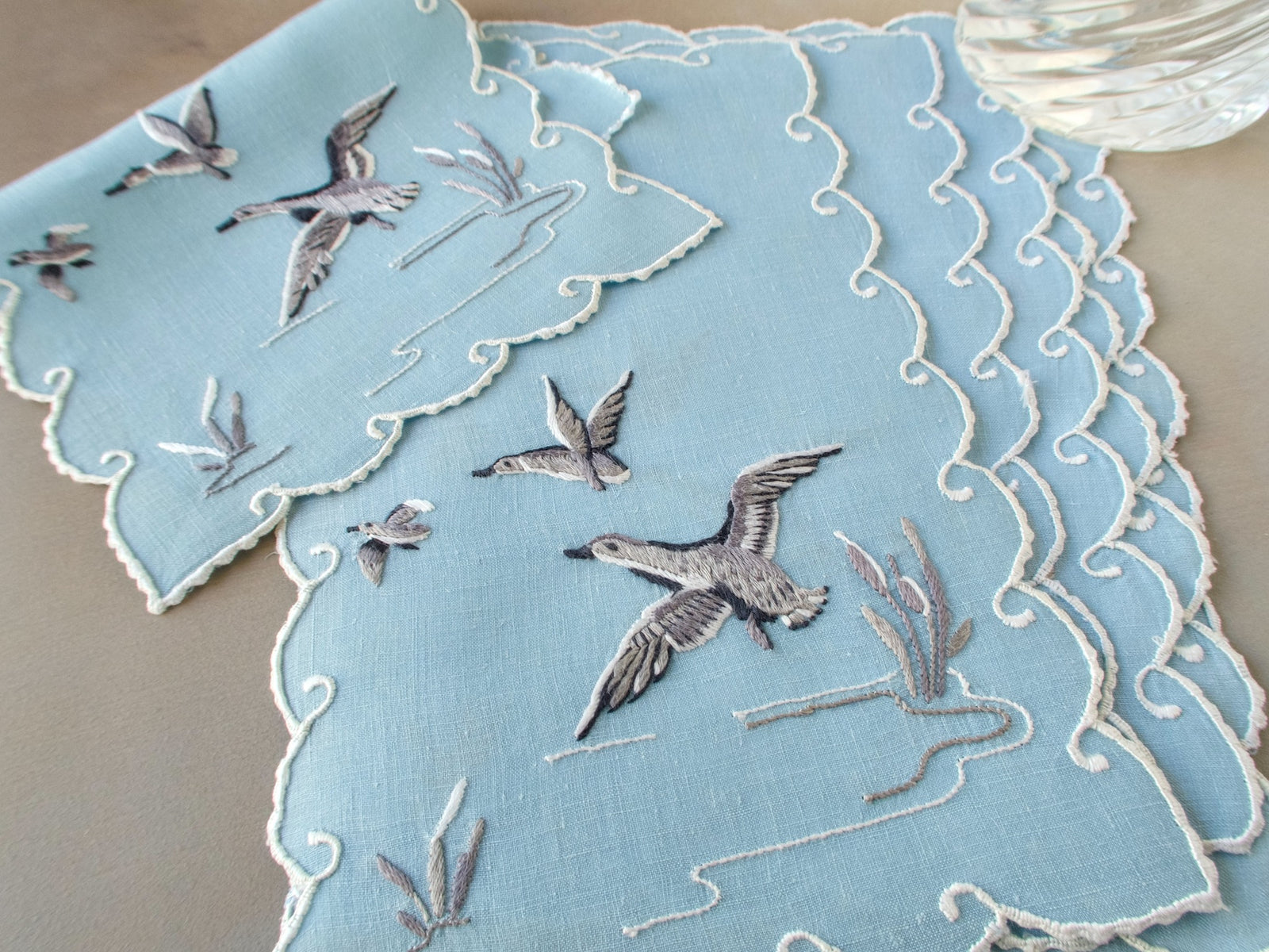 Six pale blue duck cocktail napkins arranged in a fan to show their embroidered birds and scalloped borders.