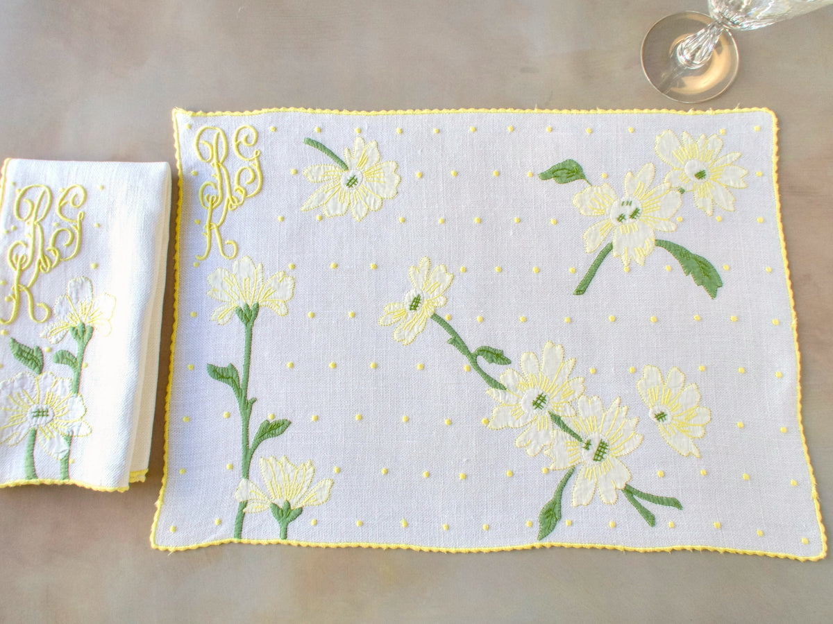 Madeira placemat and napkin laid flat, displaying white daisies, scattered dot work, and yellow cursive monogram on textured linen