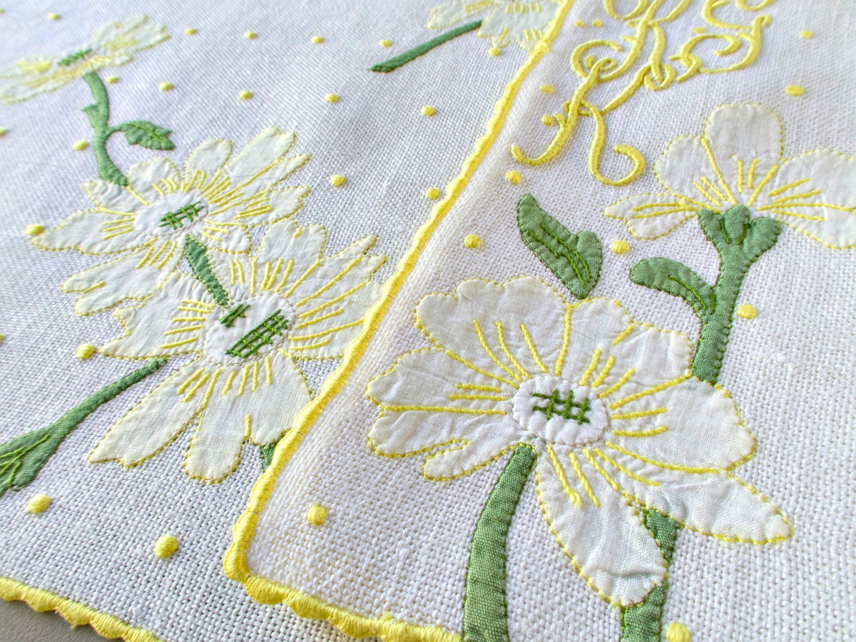 Close-up of white daisy embroidery with green leaves and yellow caseado bastido border on textured white linen, with cursive yellow monogram above