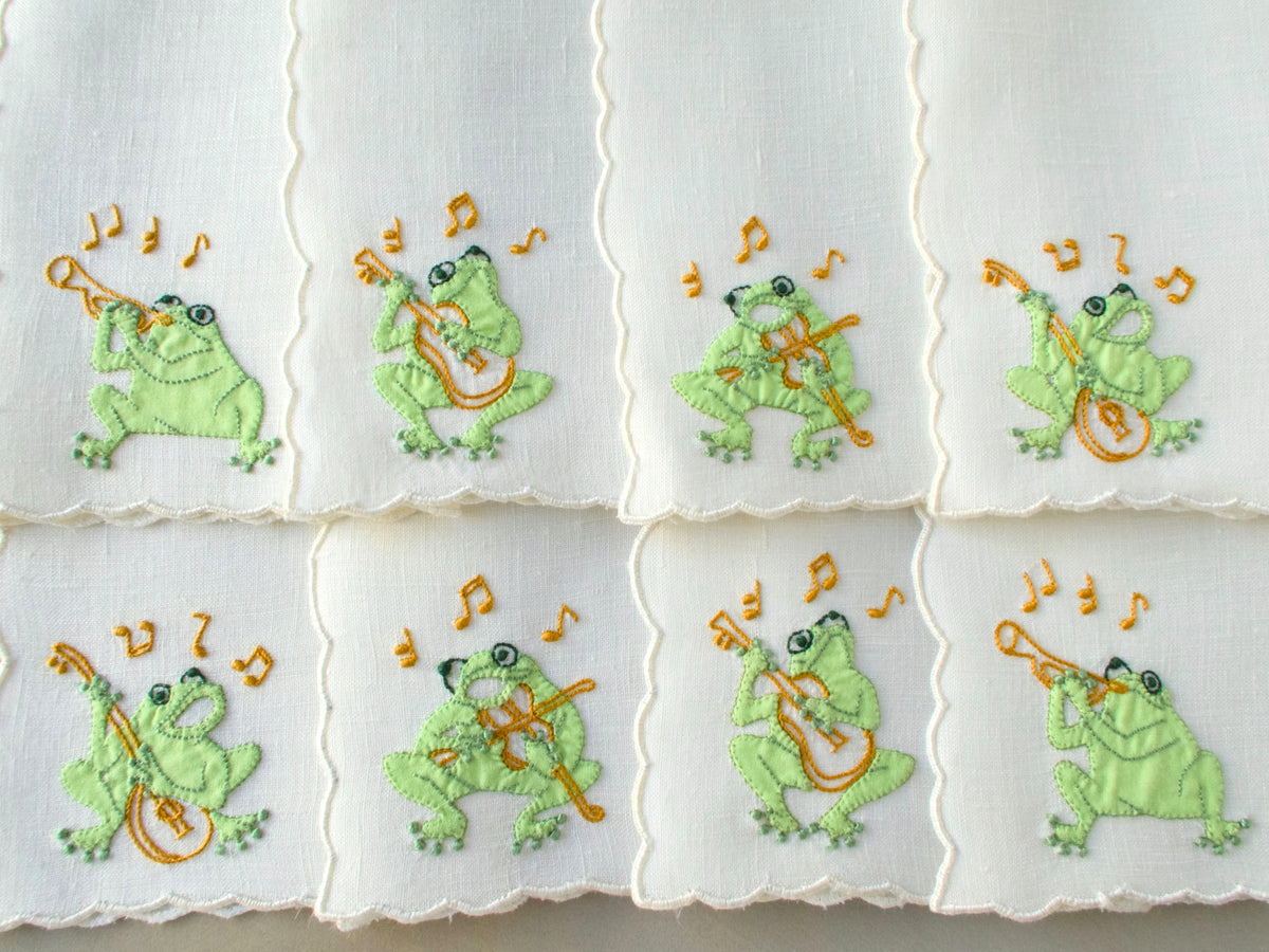 Set of eight vintage Madeira hand embroidered cocktail napkins laid out, each embroidered with a dimensional musical frog in green and gold on white linen