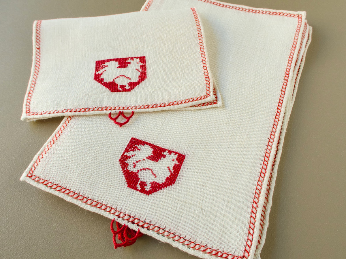 Stack of vintage Italian Assisi cocktail napkins on semi-sheer linen with a rooster in a crest, red edging and a lace embellishment